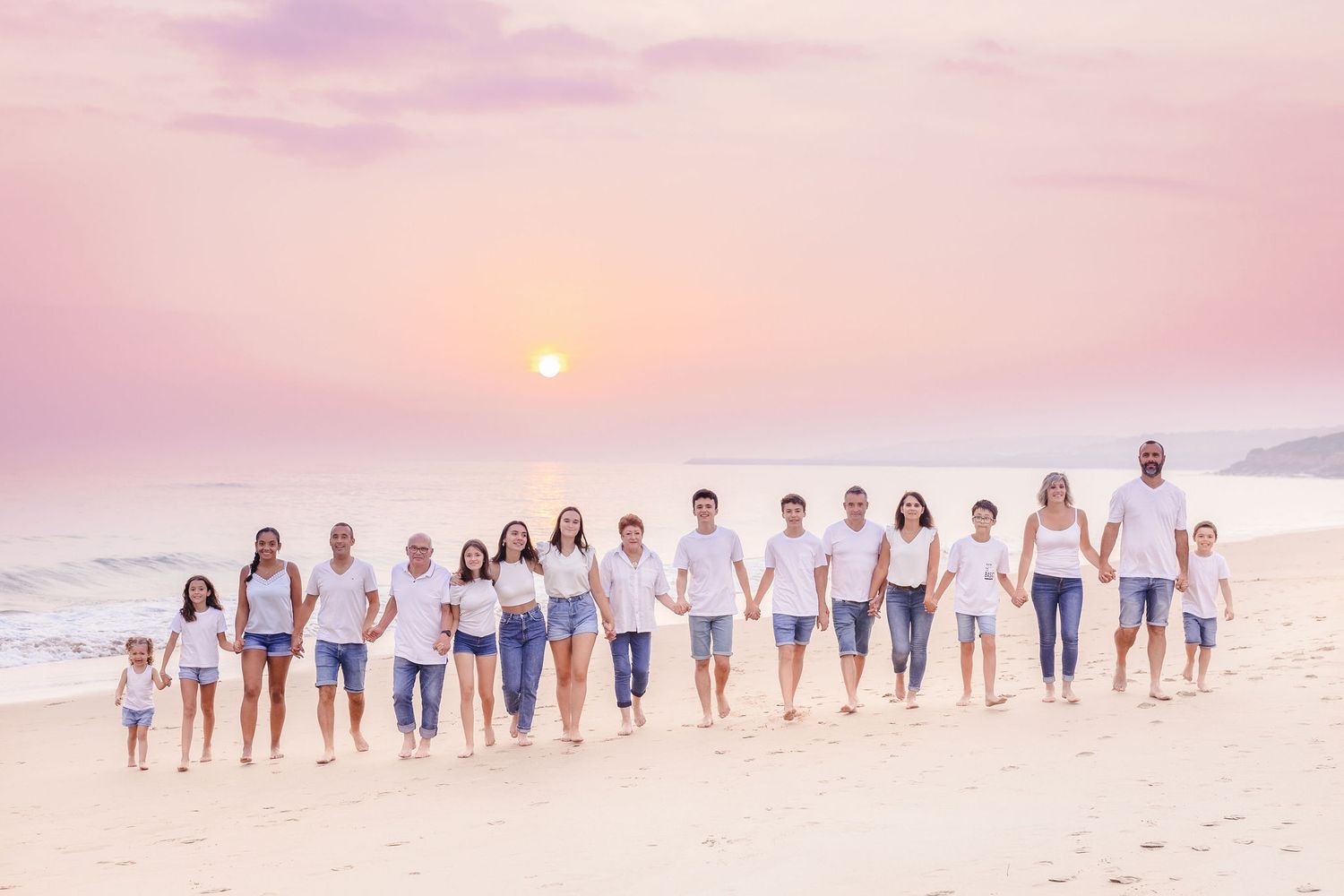 Family members hold hands in a line while walking along beach at sunset in coordinated outfits.