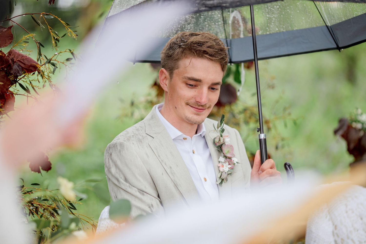 A groom in a light beige suit smiles under a clear umbrella during a rainy outdoor wedding ceremony.