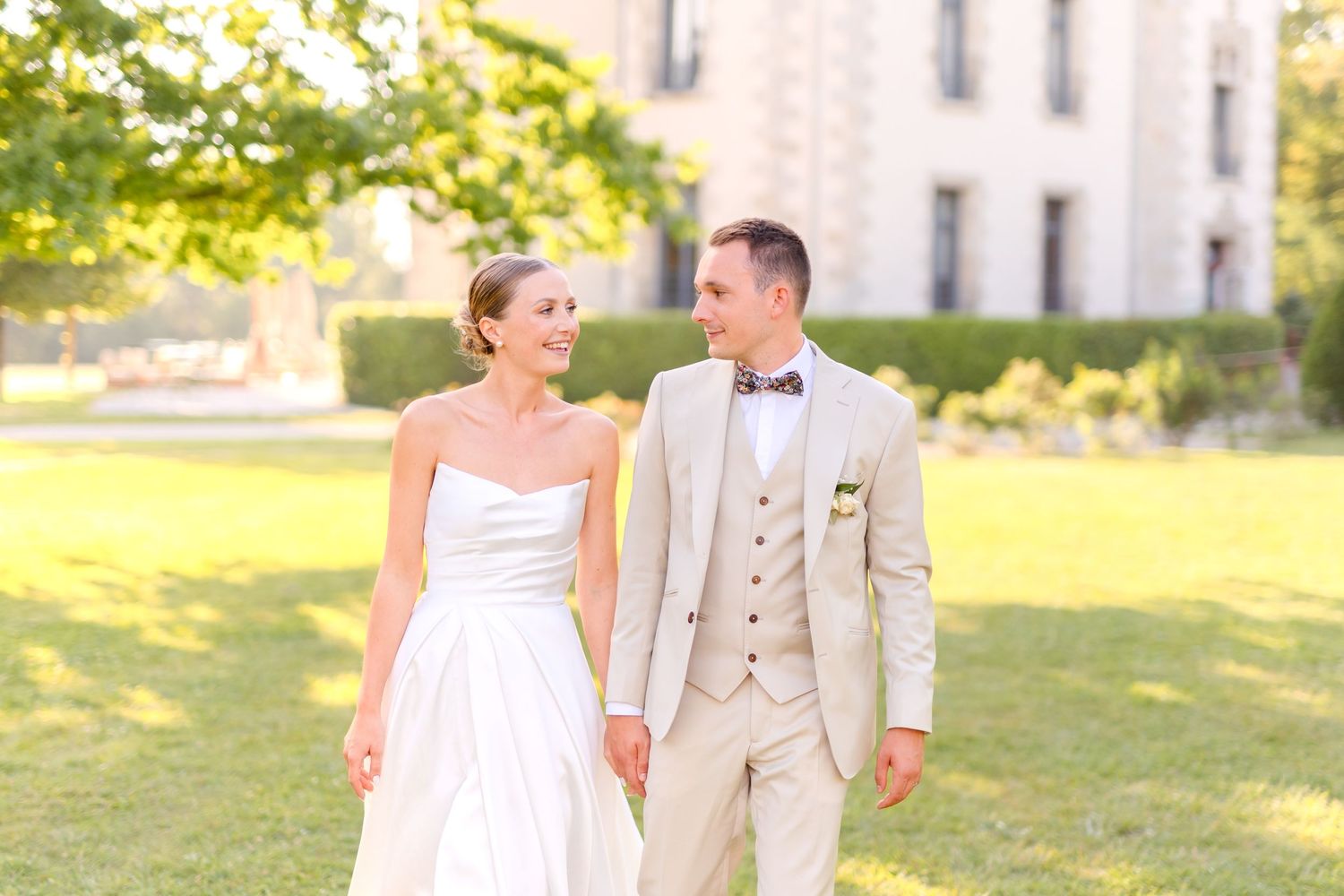 A wedding couple strolls across a sunny lawn in front of a stately chateau.