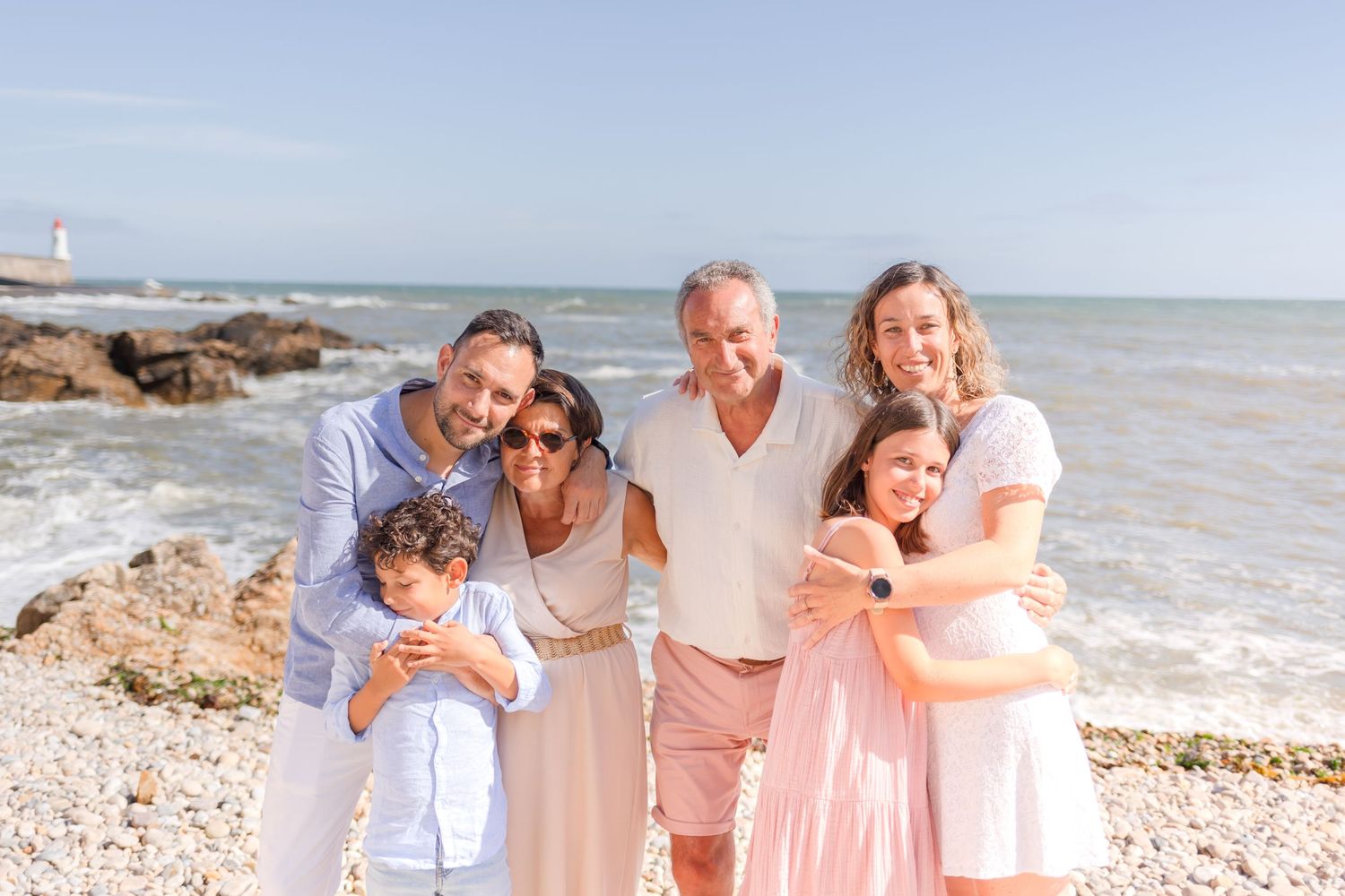 A multi-generational family group poses together on a sunny beach with waves crashing behind them.