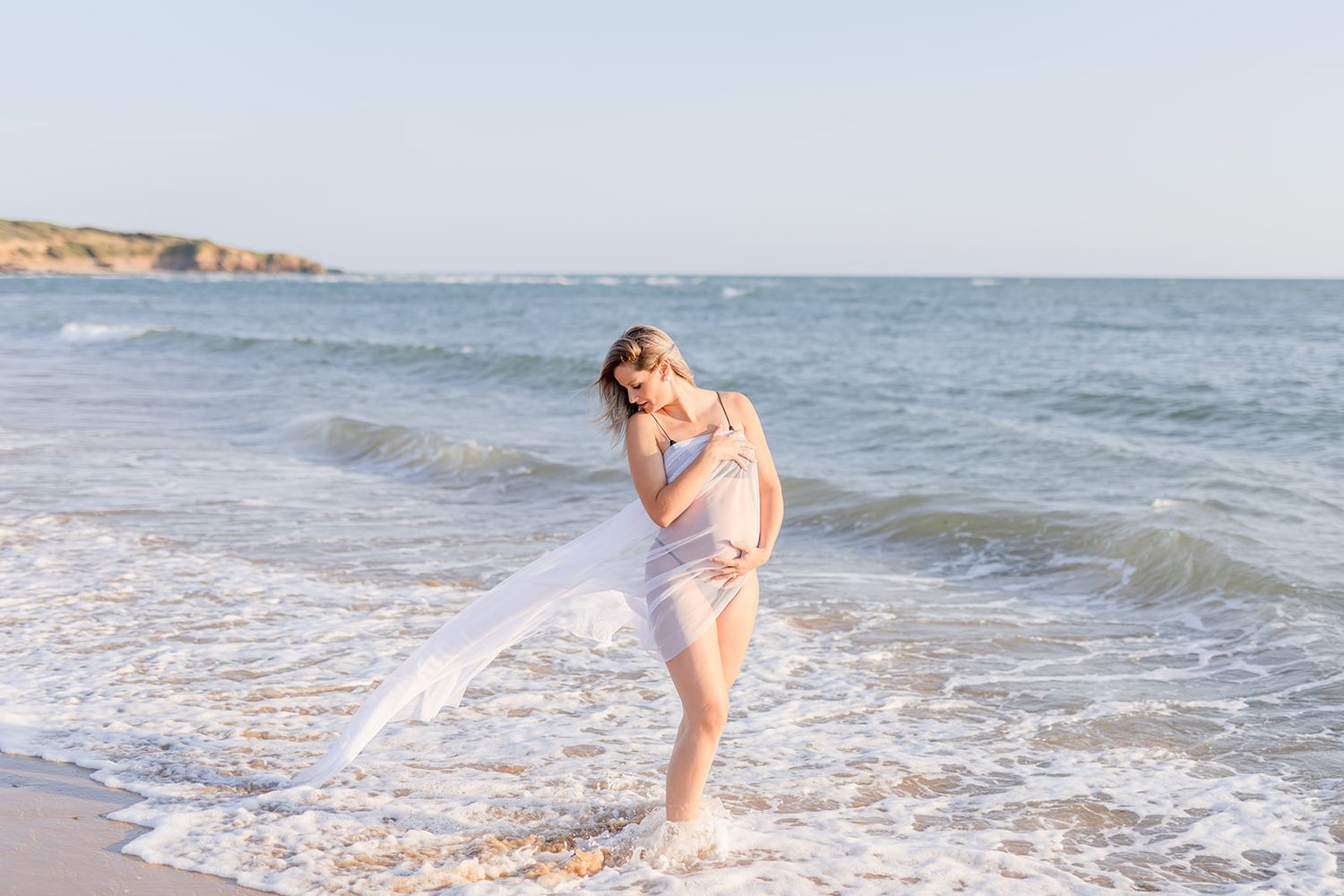 Woman in light pink swimsuit walking along ocean shoreline at sunset with waves lapping at her feet.