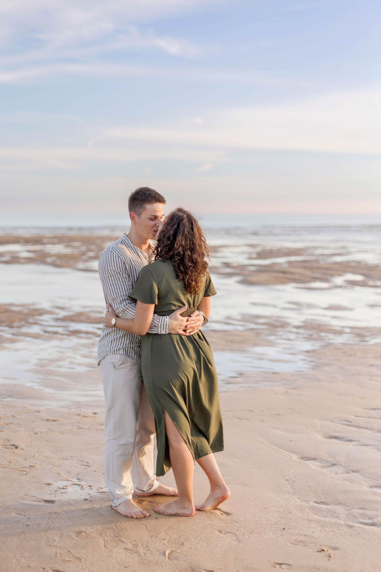 A couple in casual attire embraces while standing barefoot on a sandy beach.