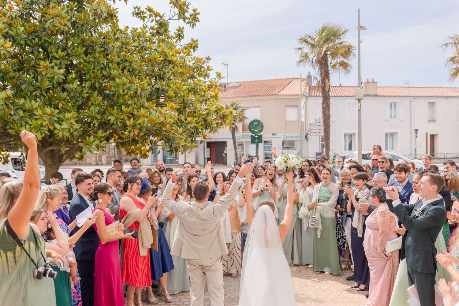 A celebrating crowd of wedding guests in colorful attire raise their hands and cheer at an outdoor ceremony.