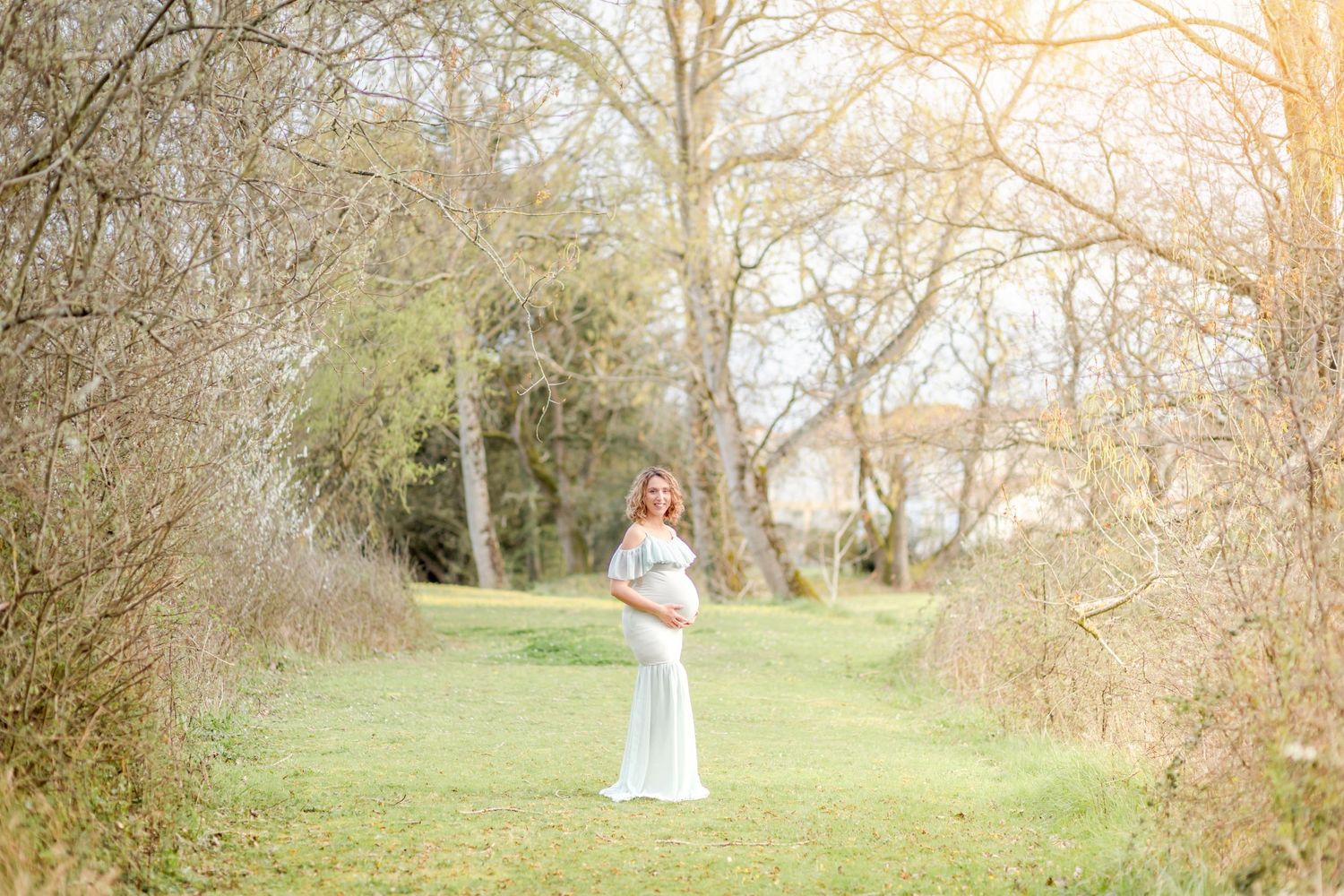 A full-length maternity photograph captured in a serene garden path with golden sunlight.