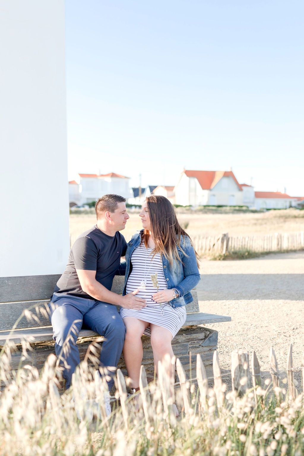 Couple sitting on bench near lighthouse with coastal houses in background.