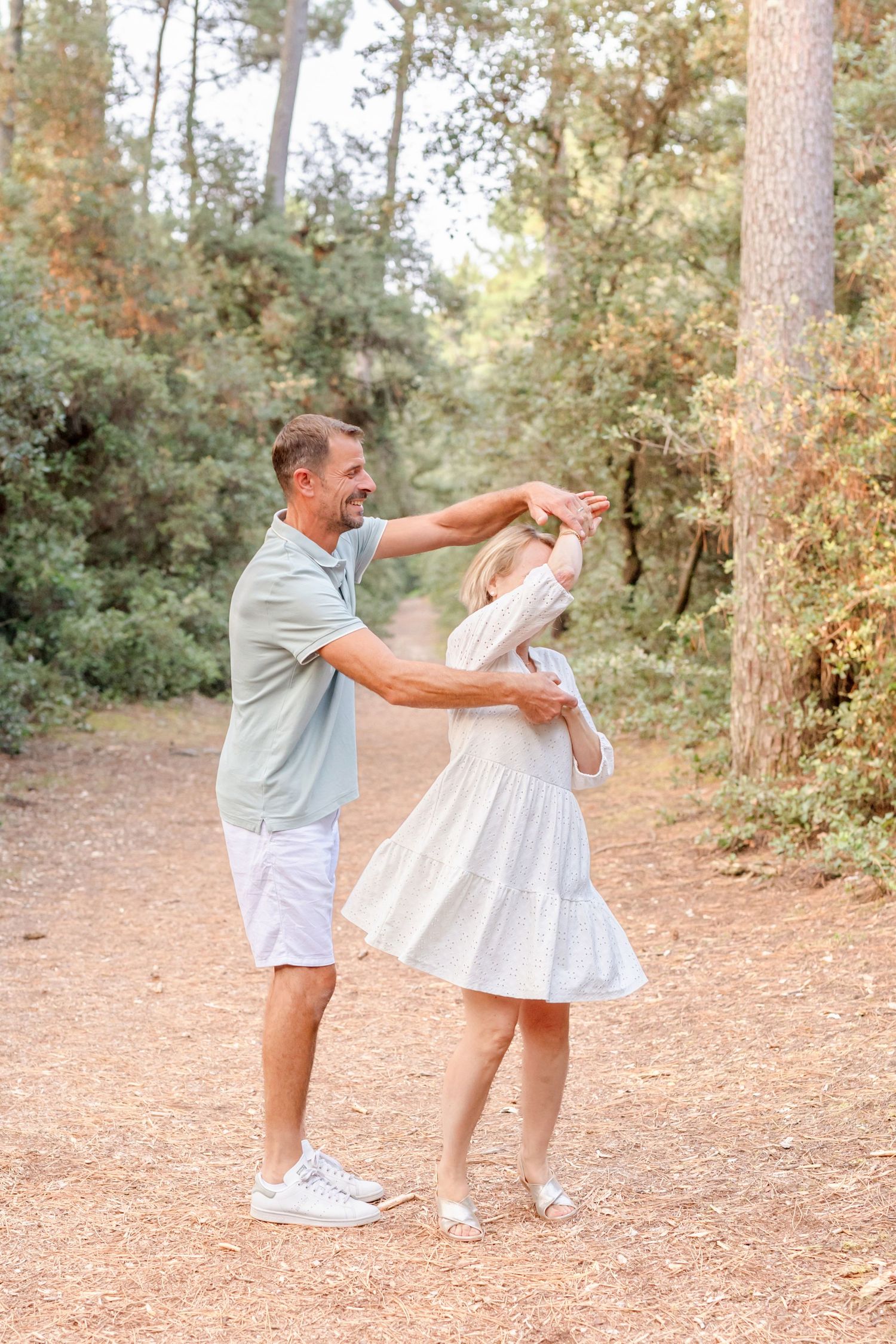 Two people in light summer outfits dance playfully on a forest path.