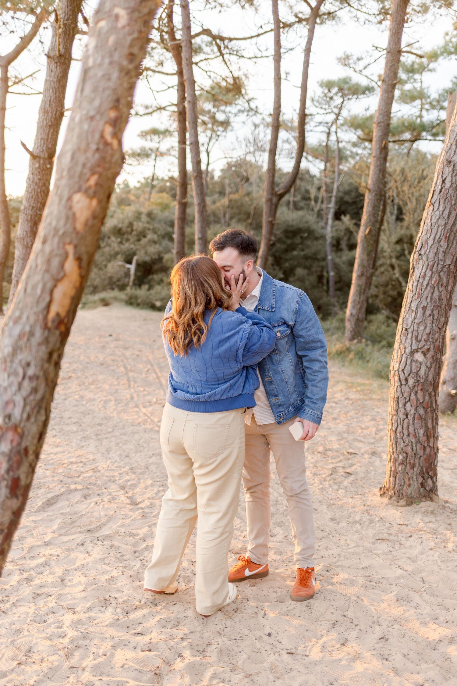 A couple in casual outfits shares an intimate moment on a forest path at golden hour.