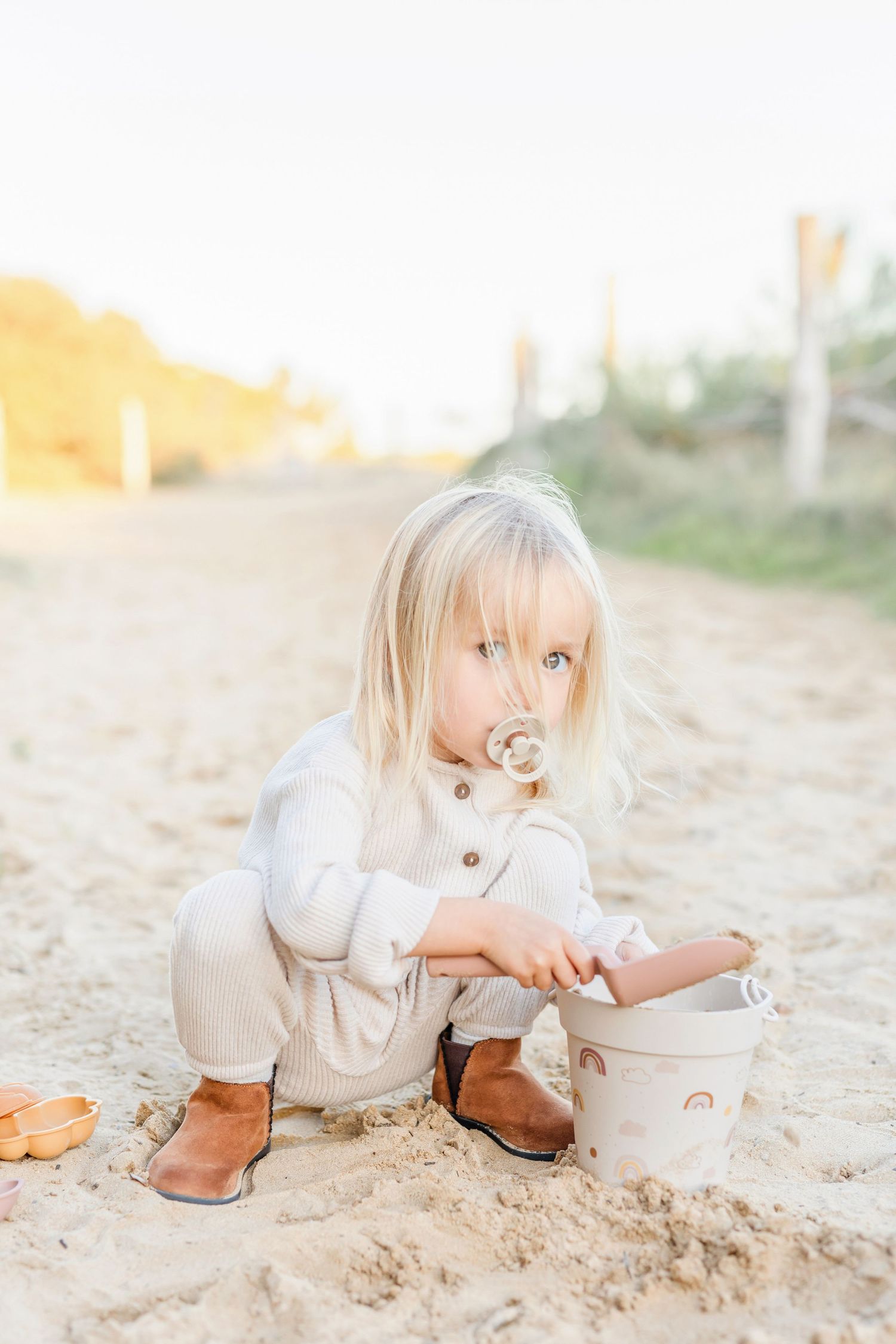 Small child in beige outfit sits on sand while playing with decorated bucket.