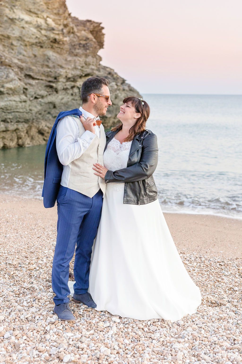 A joyful wedding couple shares a laugh on a rocky beach at sunset.