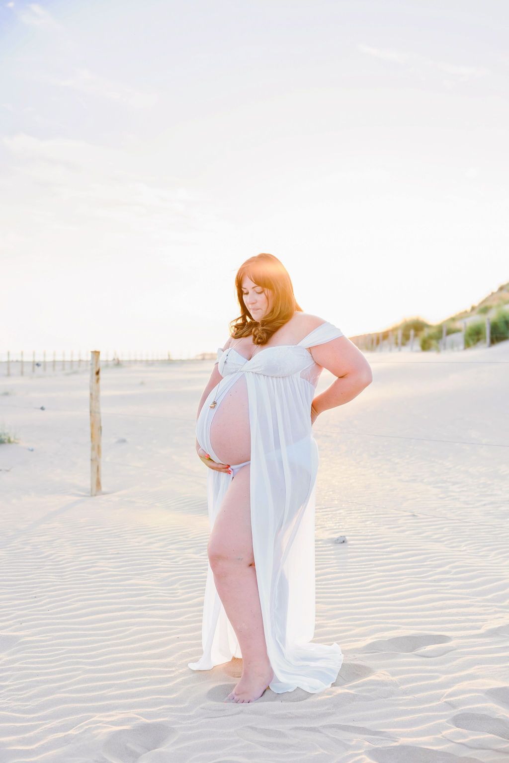 Maternity beach portrait with white flowing dress against sand dunes at golden hour.