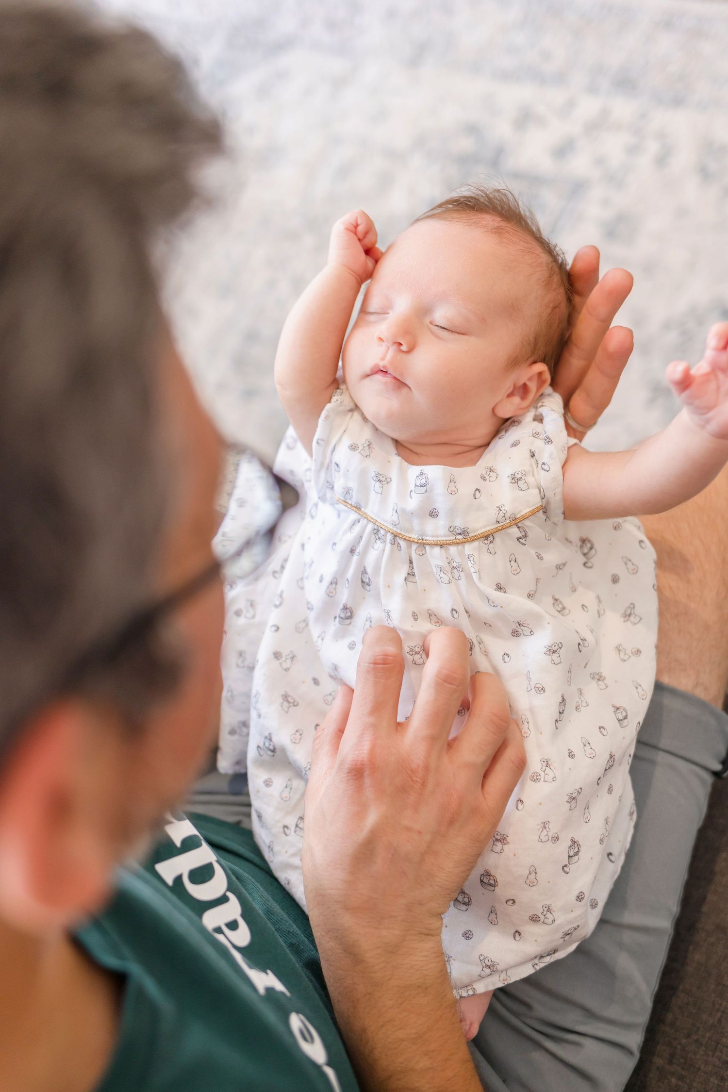A sleeping newborn baby wearing a white patterned onesie is being gently held in someone's hands.