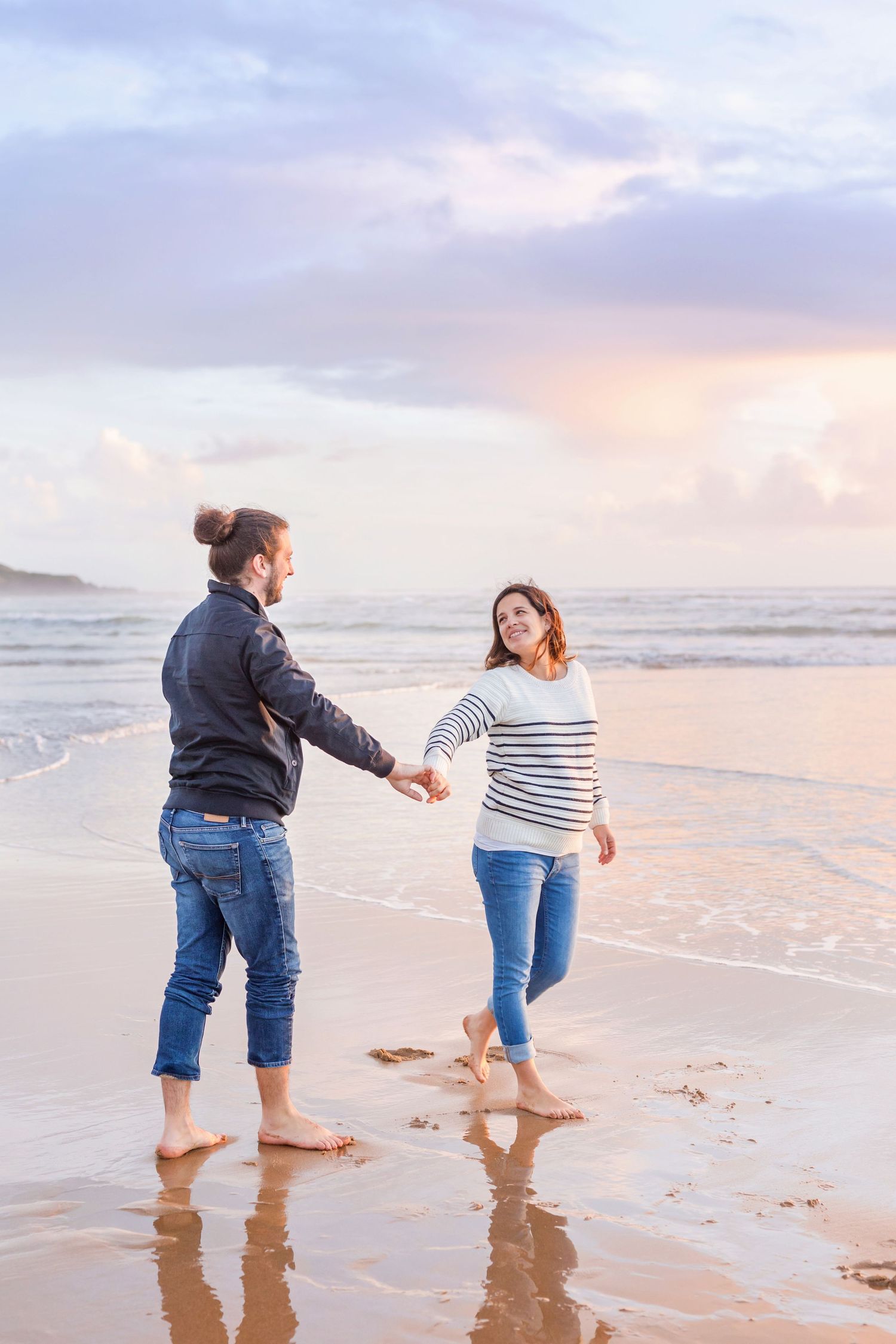 Playful couple walking along water's edge during sunset beach maternity shoot.