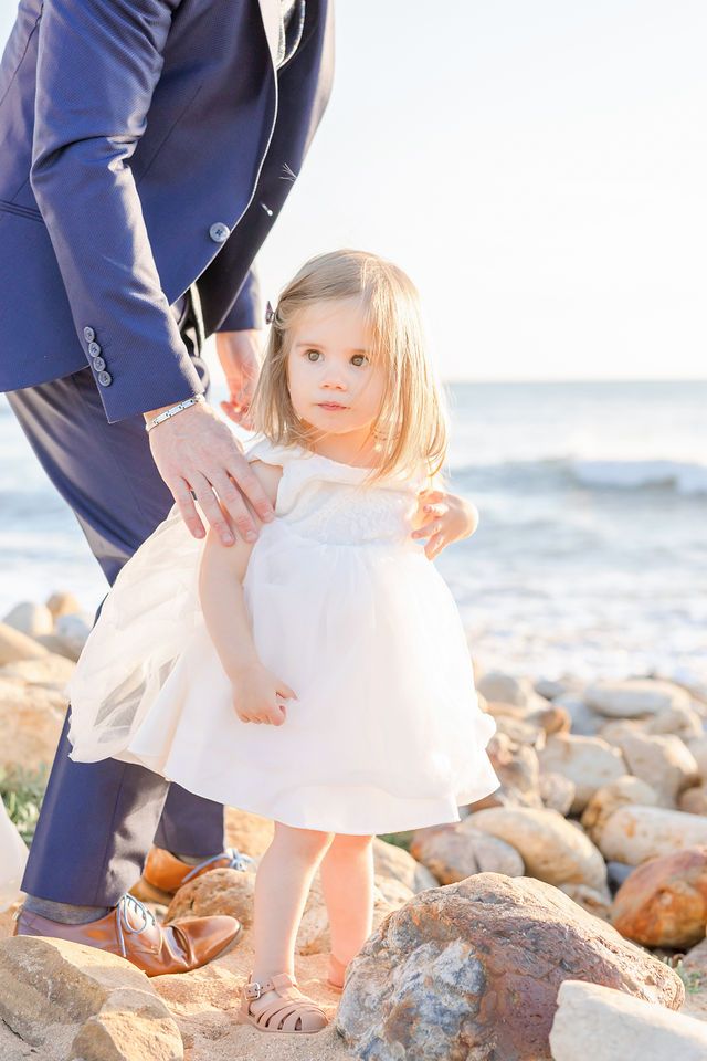 Child in white dress standing on coastal rocks with ocean waves in background.