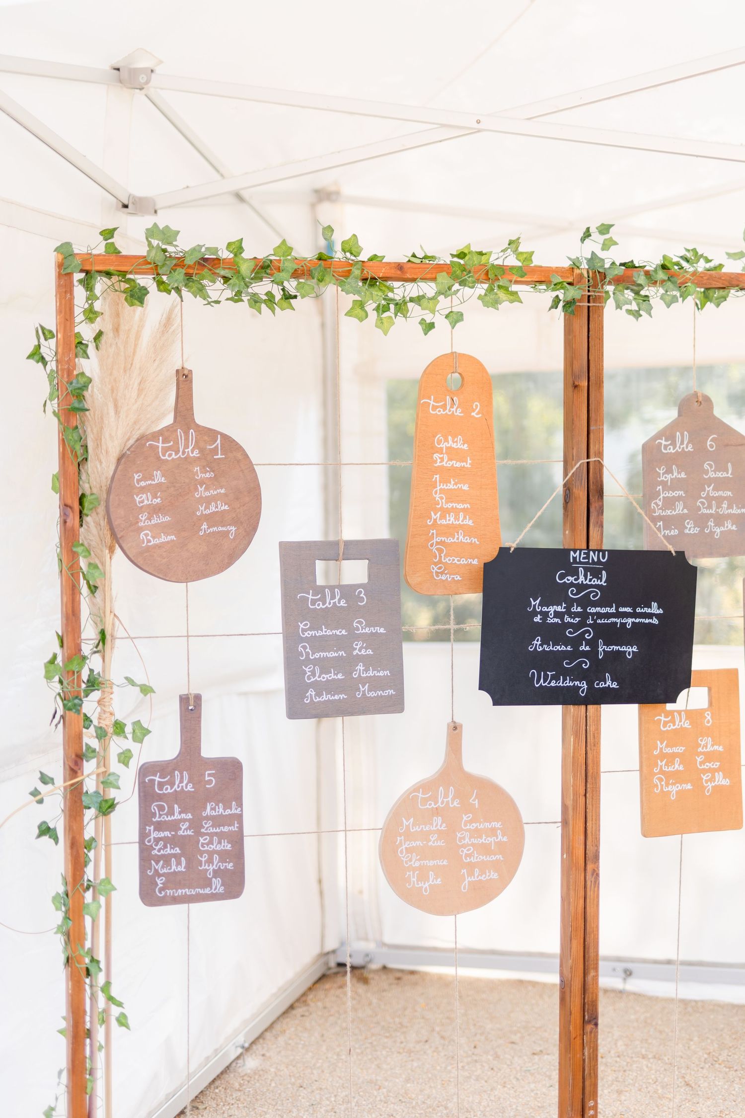 Wooden frame with hanging circular and rectangular chalkboard signs decorated with greenery for wedding seating arrangements.