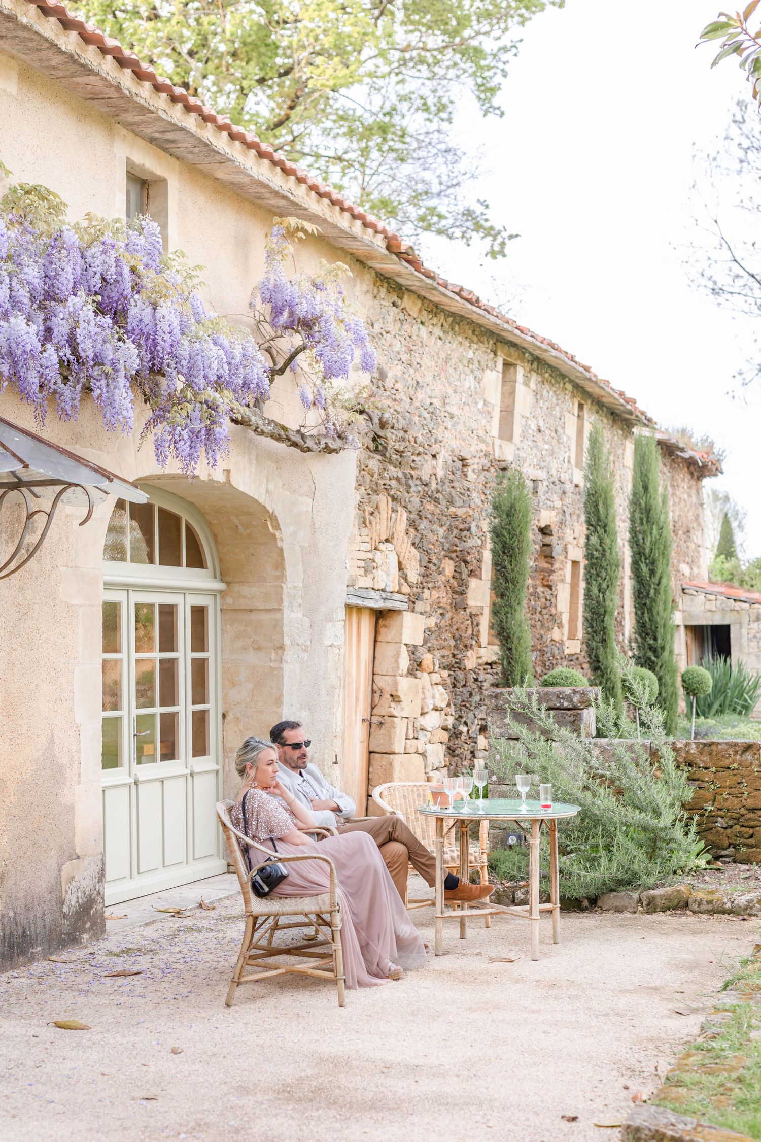 Guests relaxing in chairs under wisteria-covered facade of stone building.
