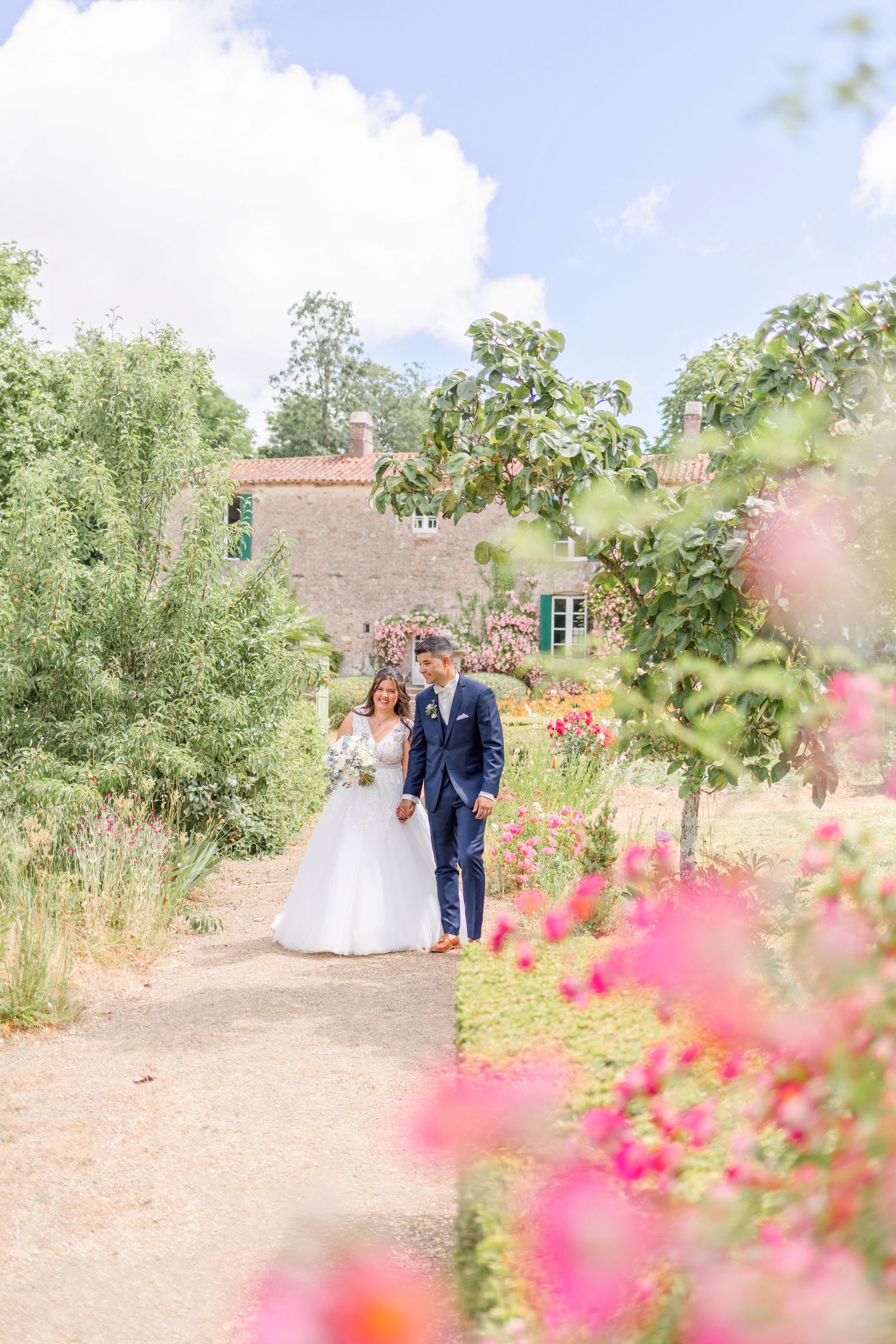 A couple in wedding attire strolls down a garden path with pink flowers and greenery along a stone building.