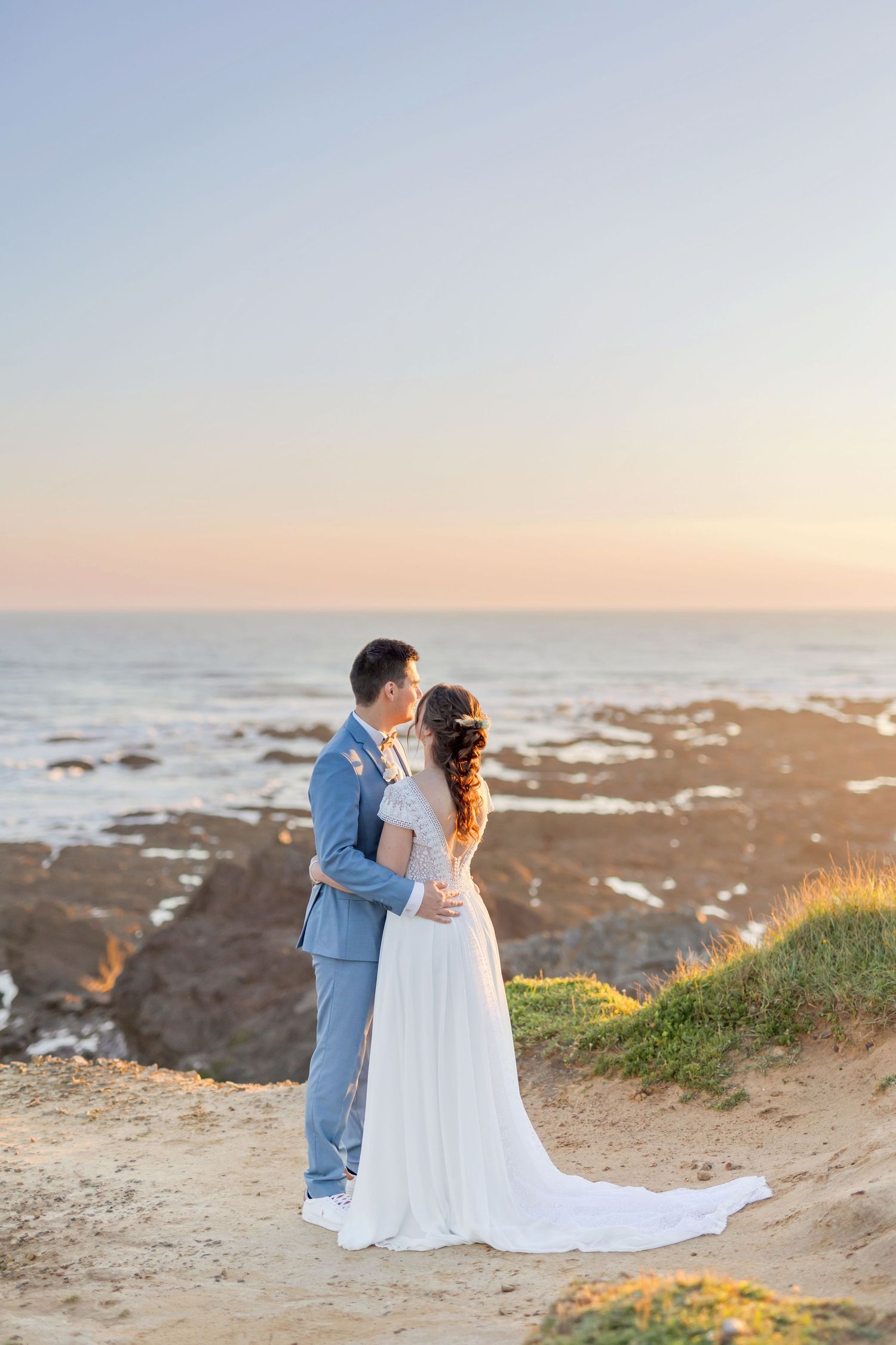 Romantic seaside wedding photo with couple embracing on coastal cliffs at sunset.