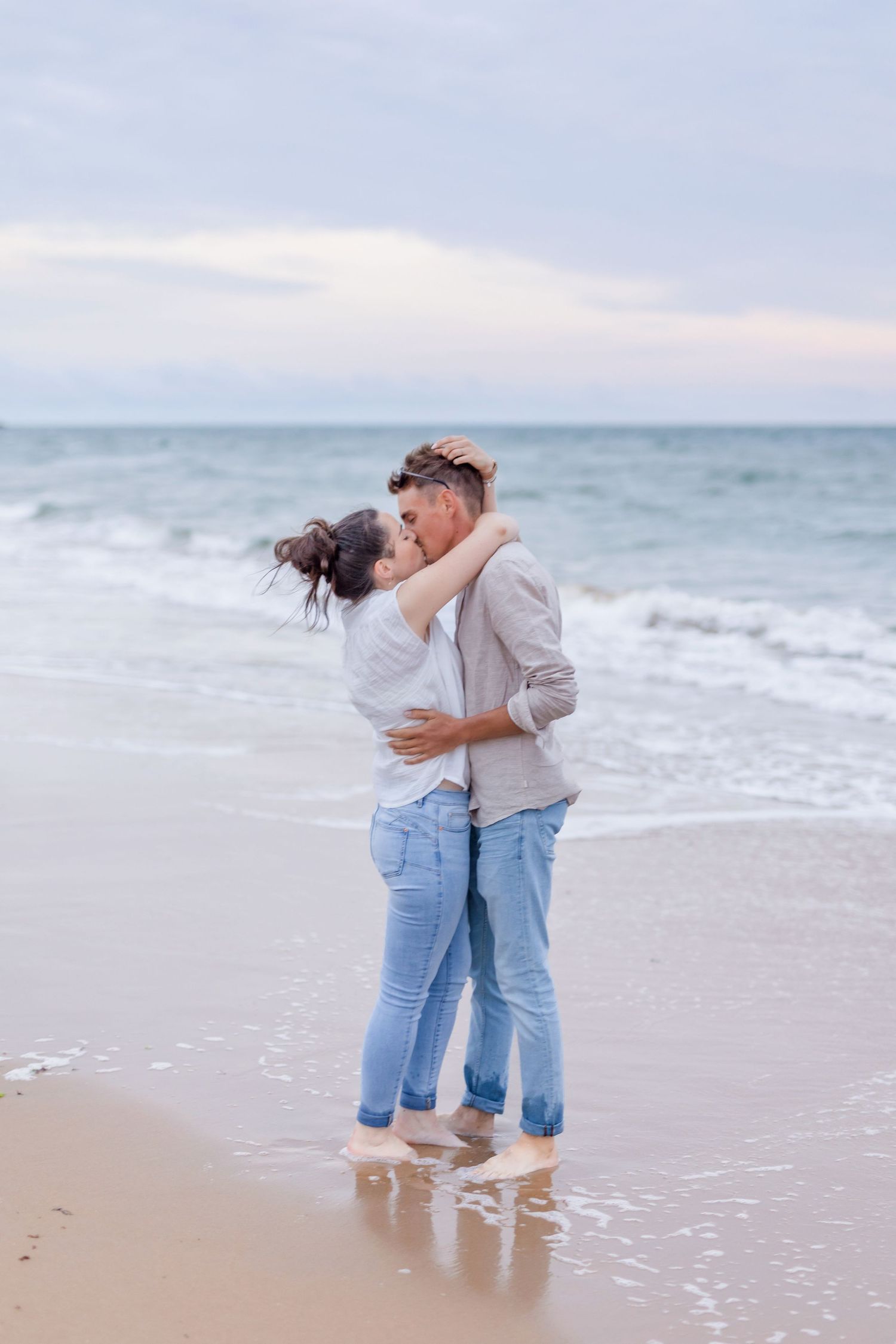 A romantic couple embraces on a serene beach at sunset with ocean waves in the background.