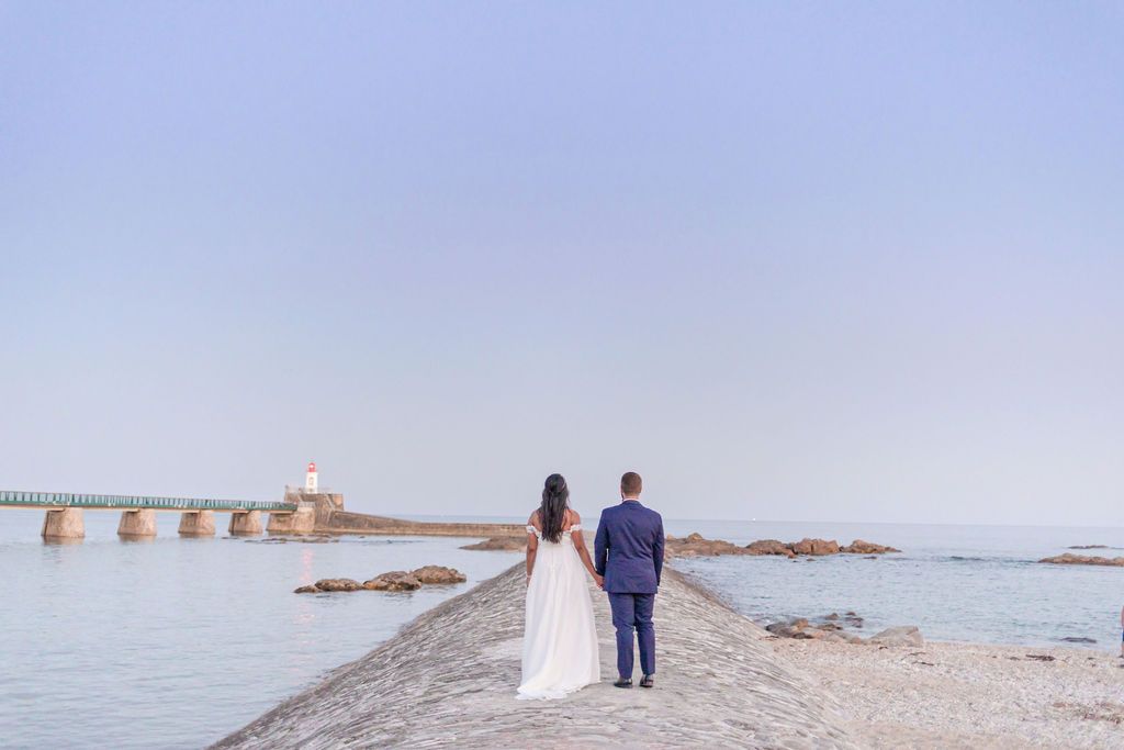 Wedding couple walks along historic stone pier with lighthouse in the distance.