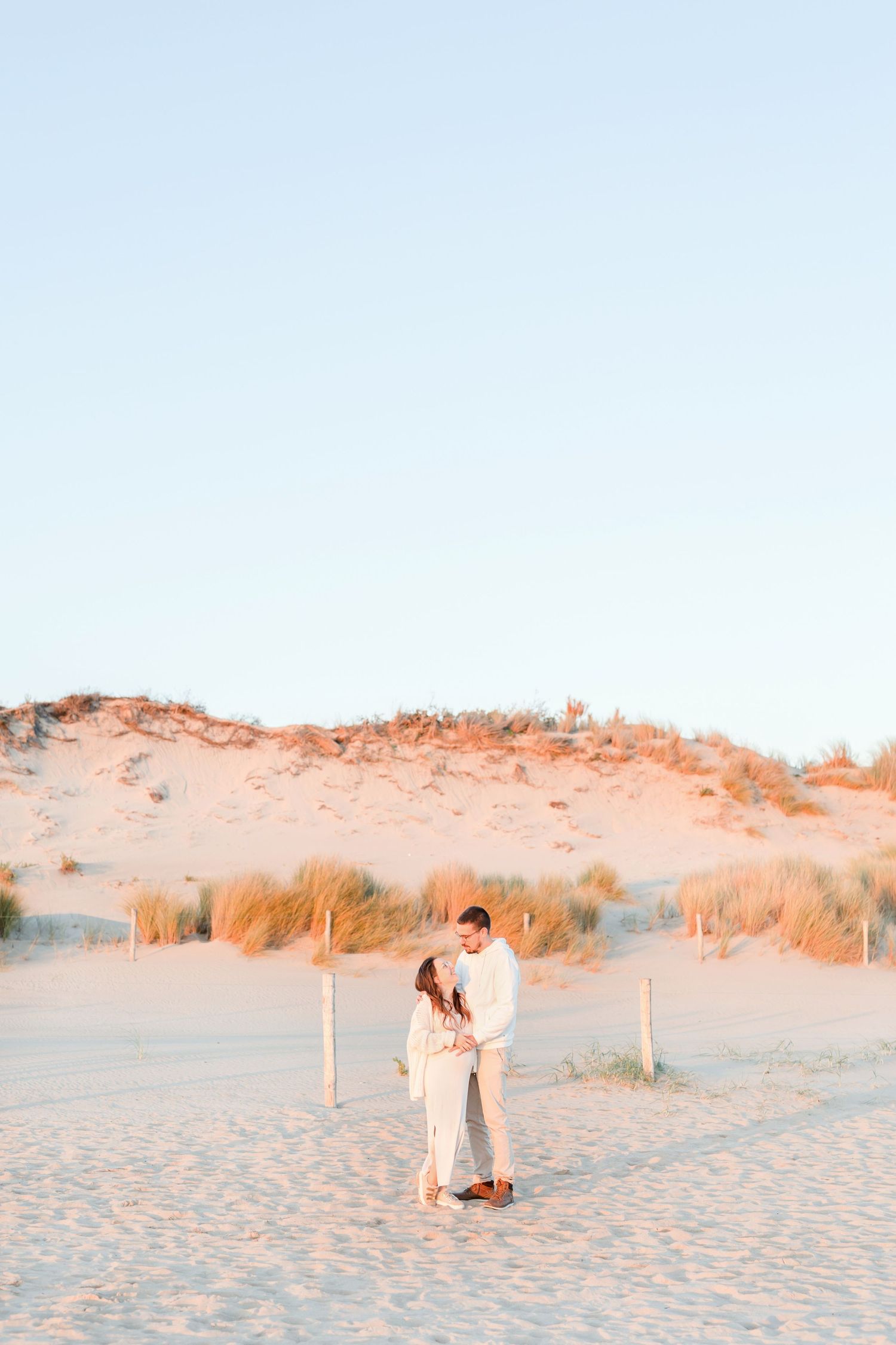 A couple embraces on a serene beach with sand dunes in the warm evening light.