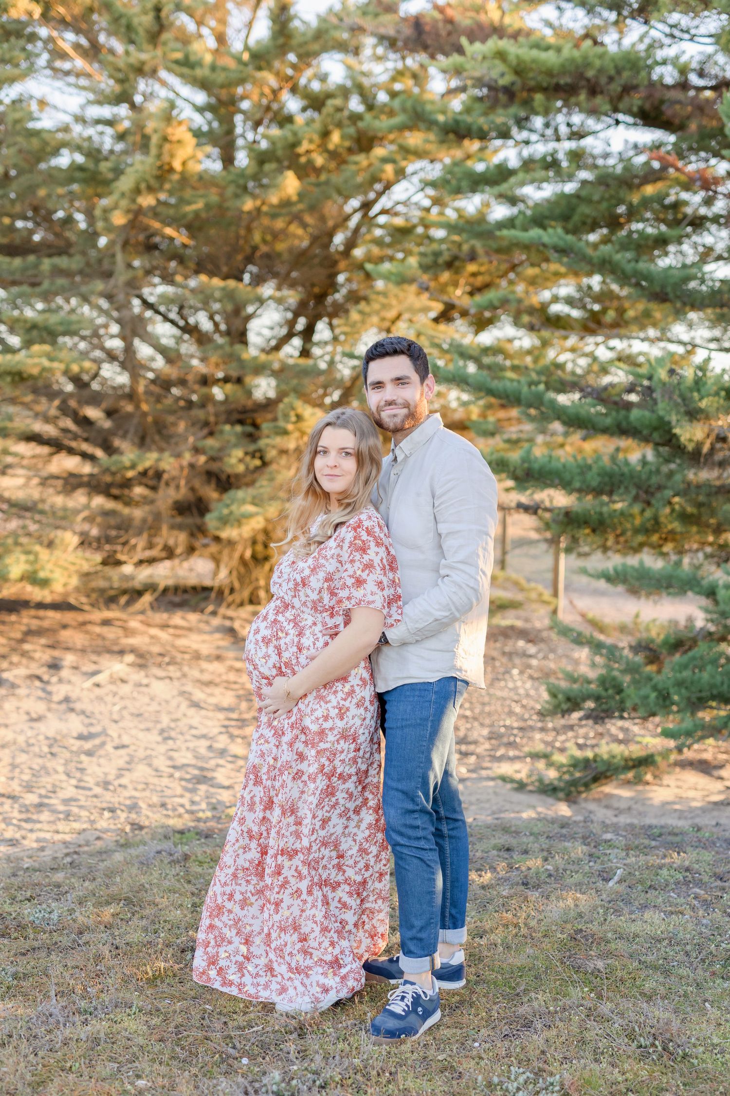 Maternity portrait of couple standing together among pine trees at sunset.