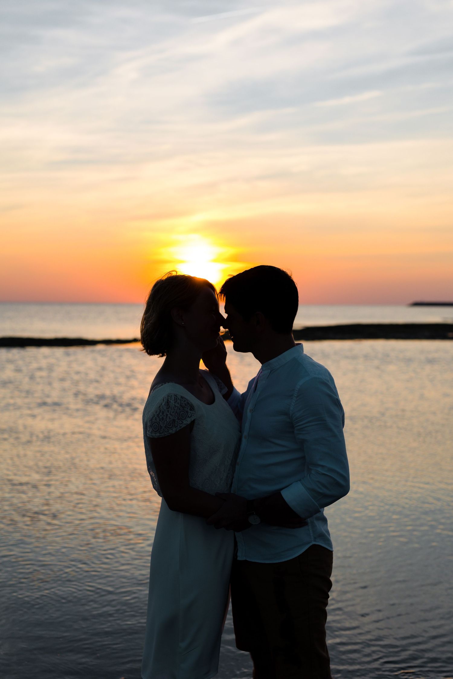 Silhouettes of a couple embrace against a dramatic orange sunset over calm ocean waters.