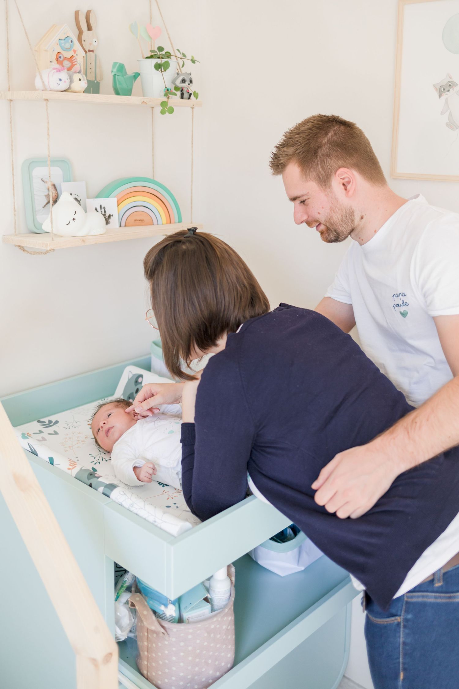 Parents changing their baby on a mint green dresser with rainbow decor on shelf above.