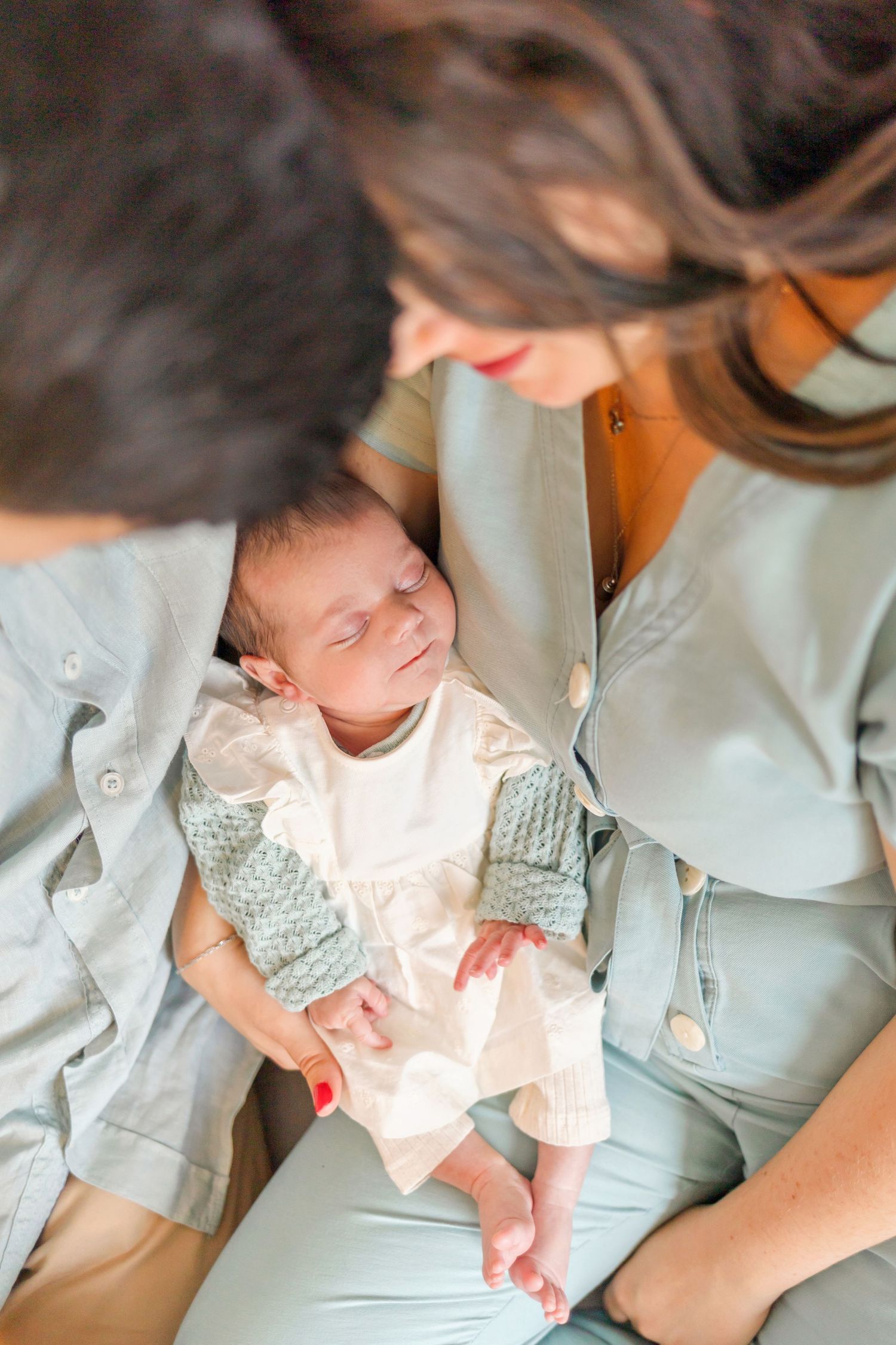Sleeping newborn in white outfit nestled between caretakers in soft lighting.