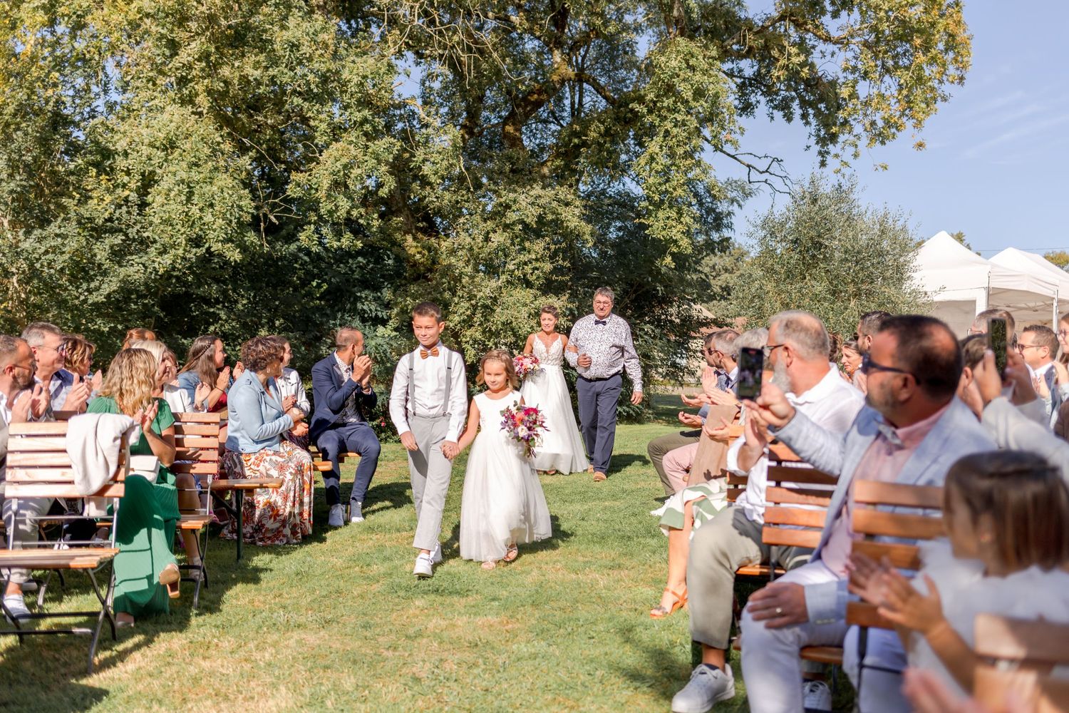 Wedding processional walking down aisle between seated guests at outdoor ceremony.