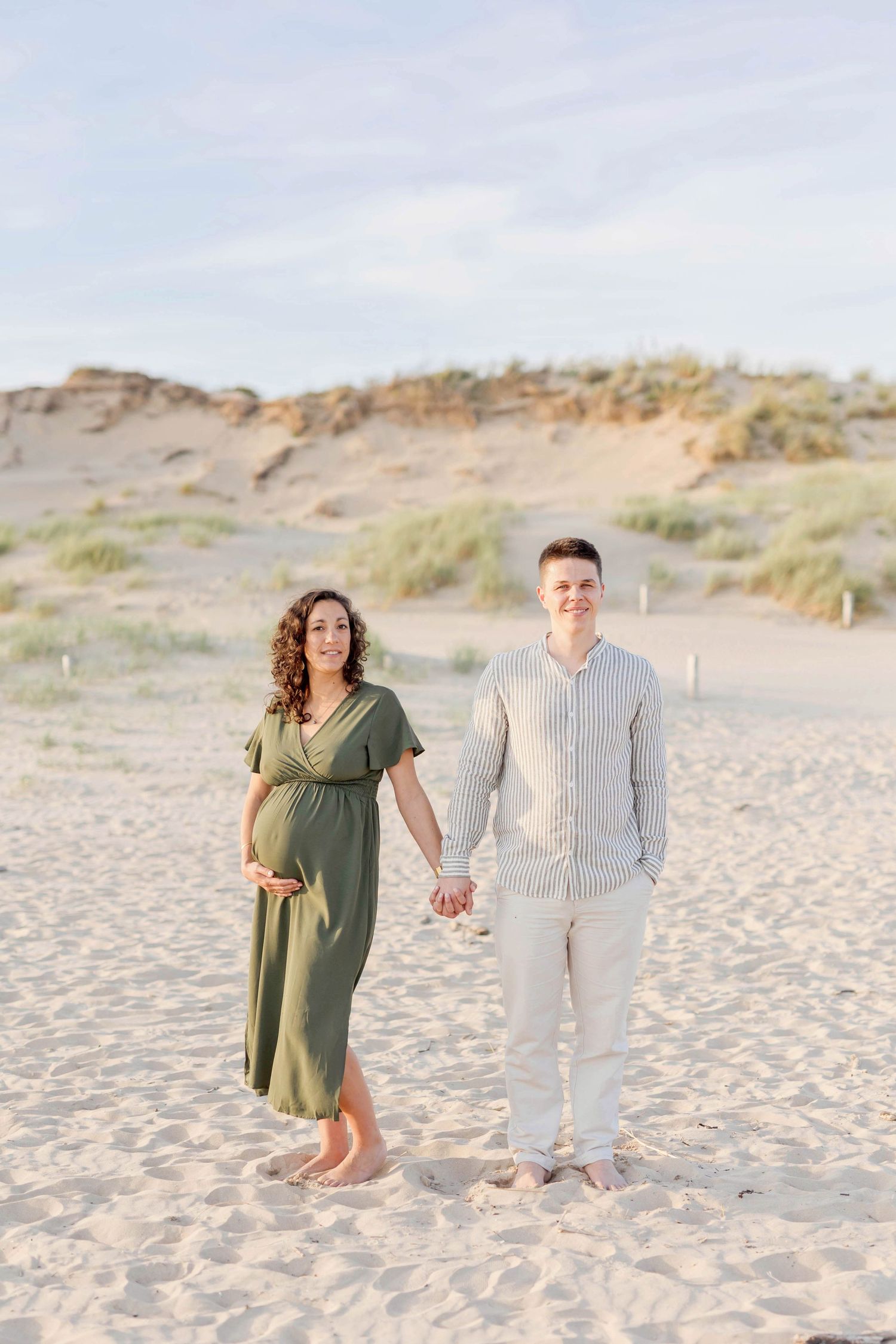 A couple stands barefoot on beach sand with coastal dunes in the background.