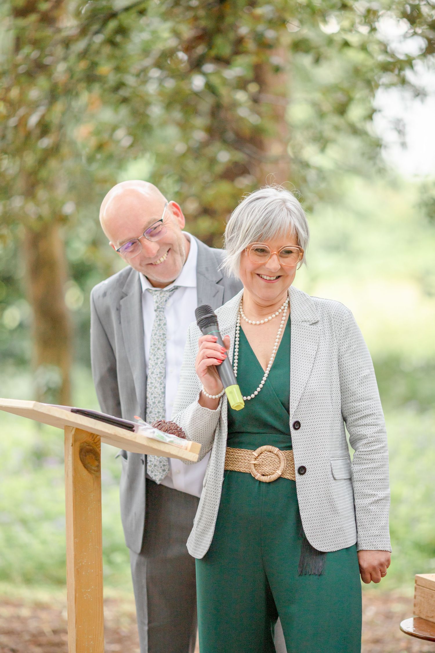 Couple in coordinating green and grey outfits present at wooden podium during outdoor ceremony.