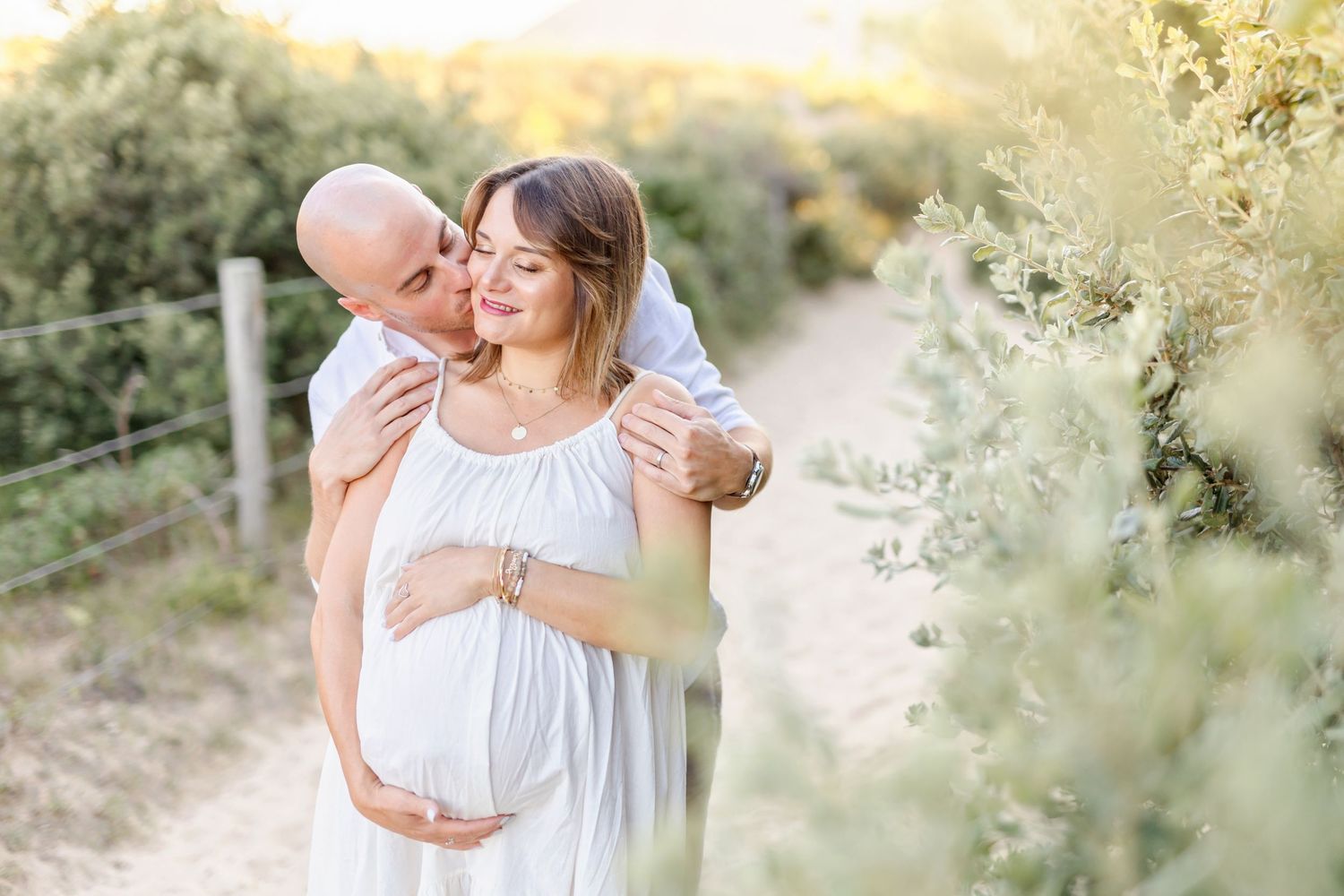 Romantic maternity photo of couple on outdoor trail with soft evening light and greenery.