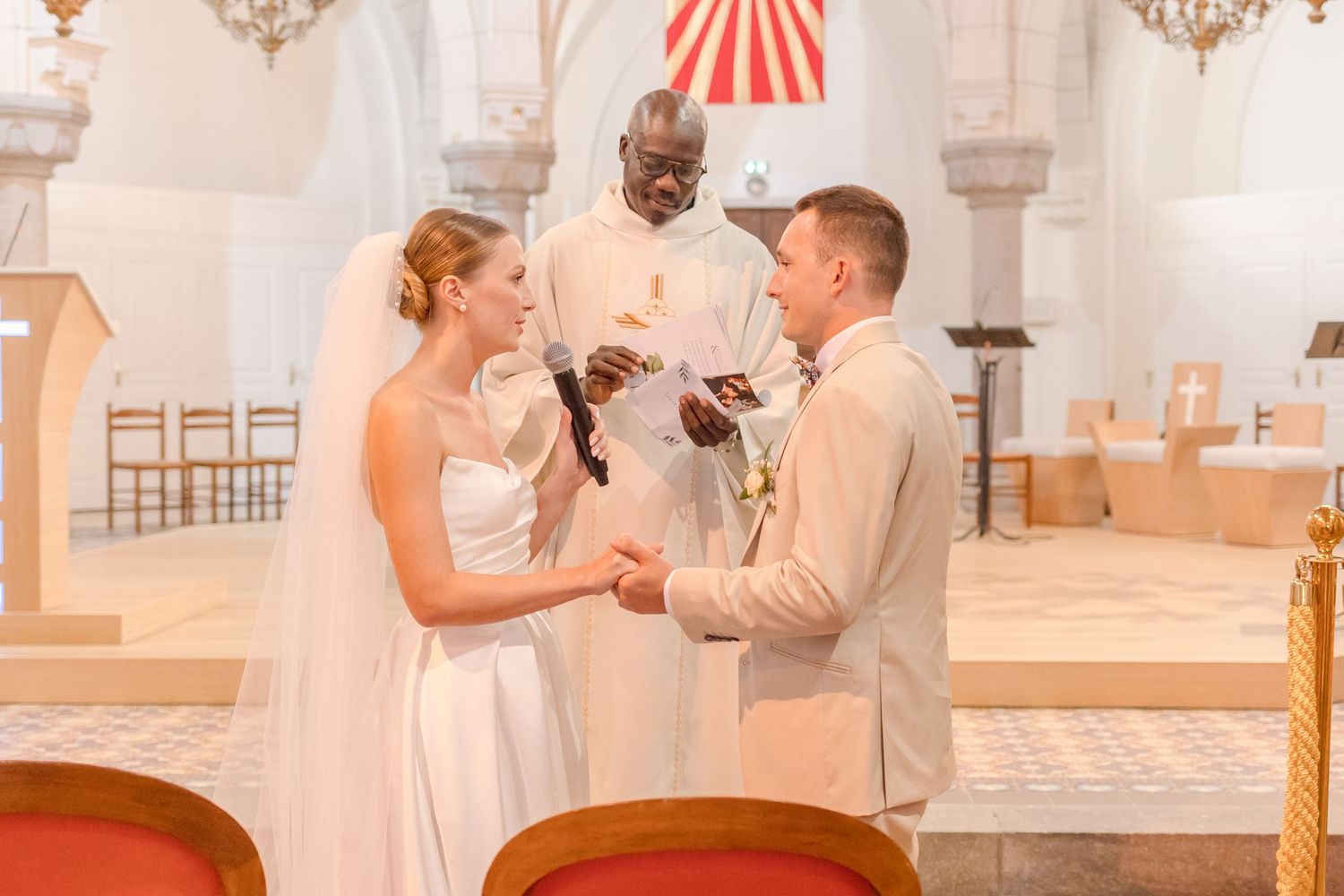 A wedding ceremony takes place in an elegant church with ornate architecture and red banners.