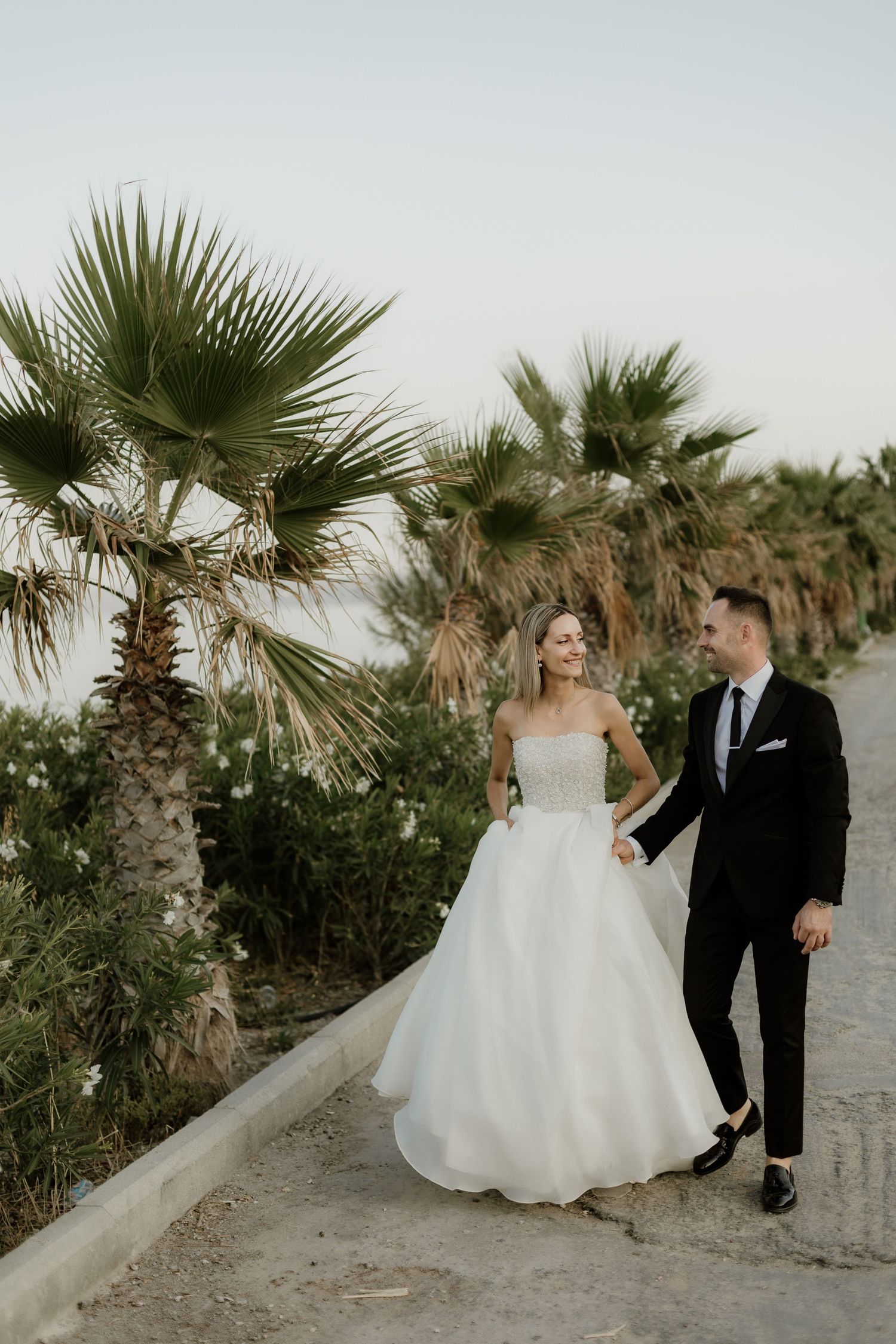 Newlyweds walking along palm tree-lined path in elegant wedding attire.