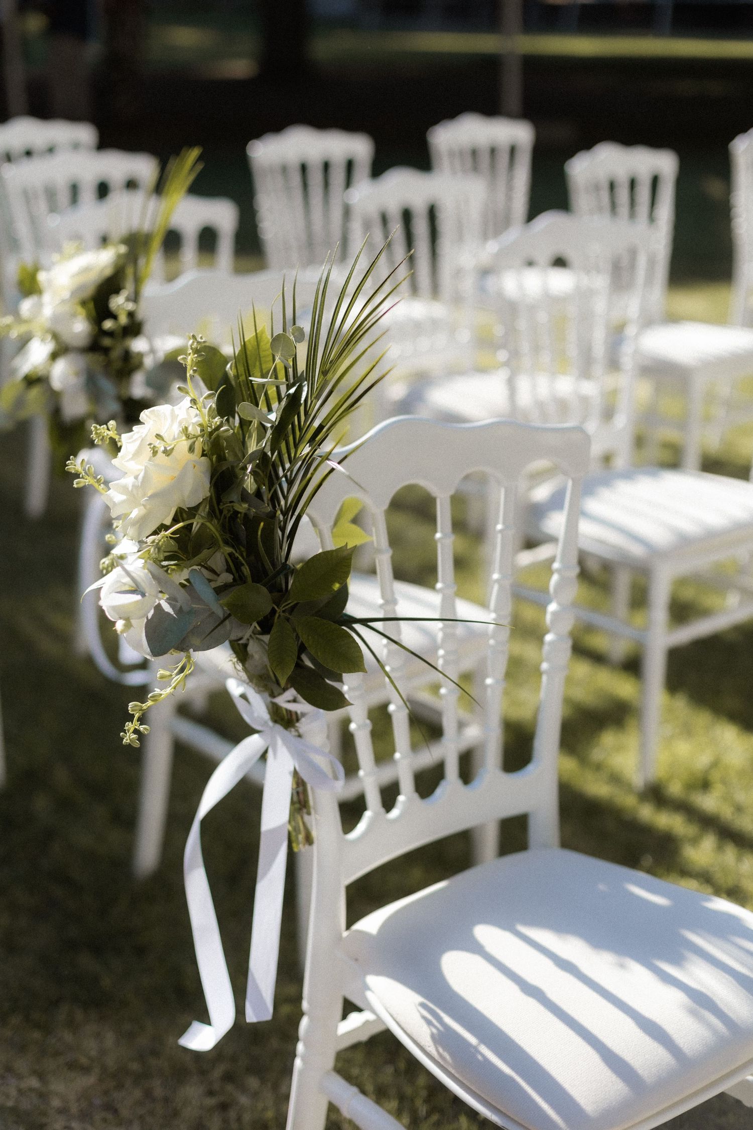 White chairs decorated with floral arrangements set up for an outdoor wedding ceremony.