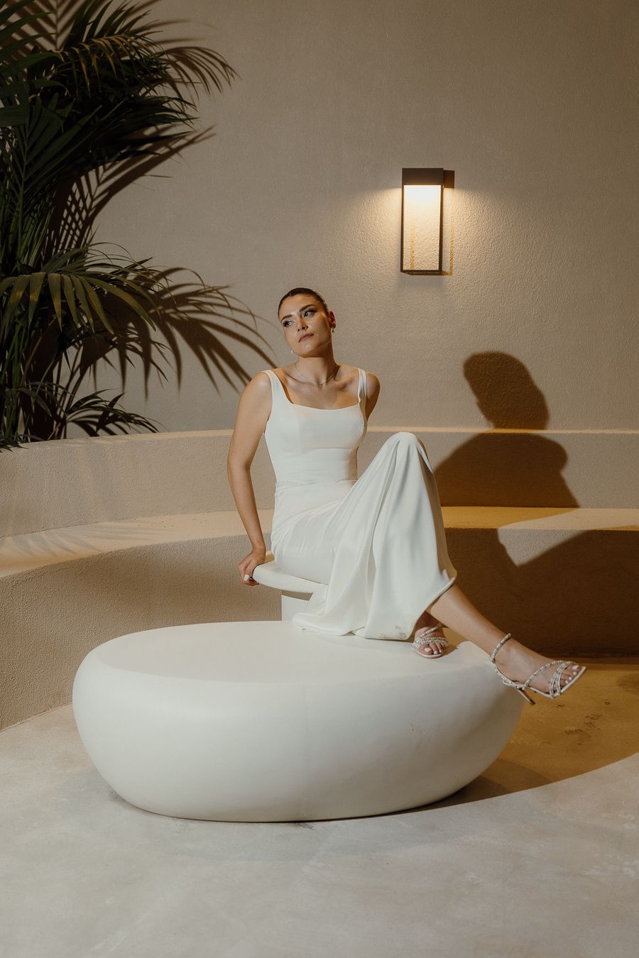 Woman in white jumpsuit sitting on a round white ottoman in an elegant, warmly lit interior with tropical plants.