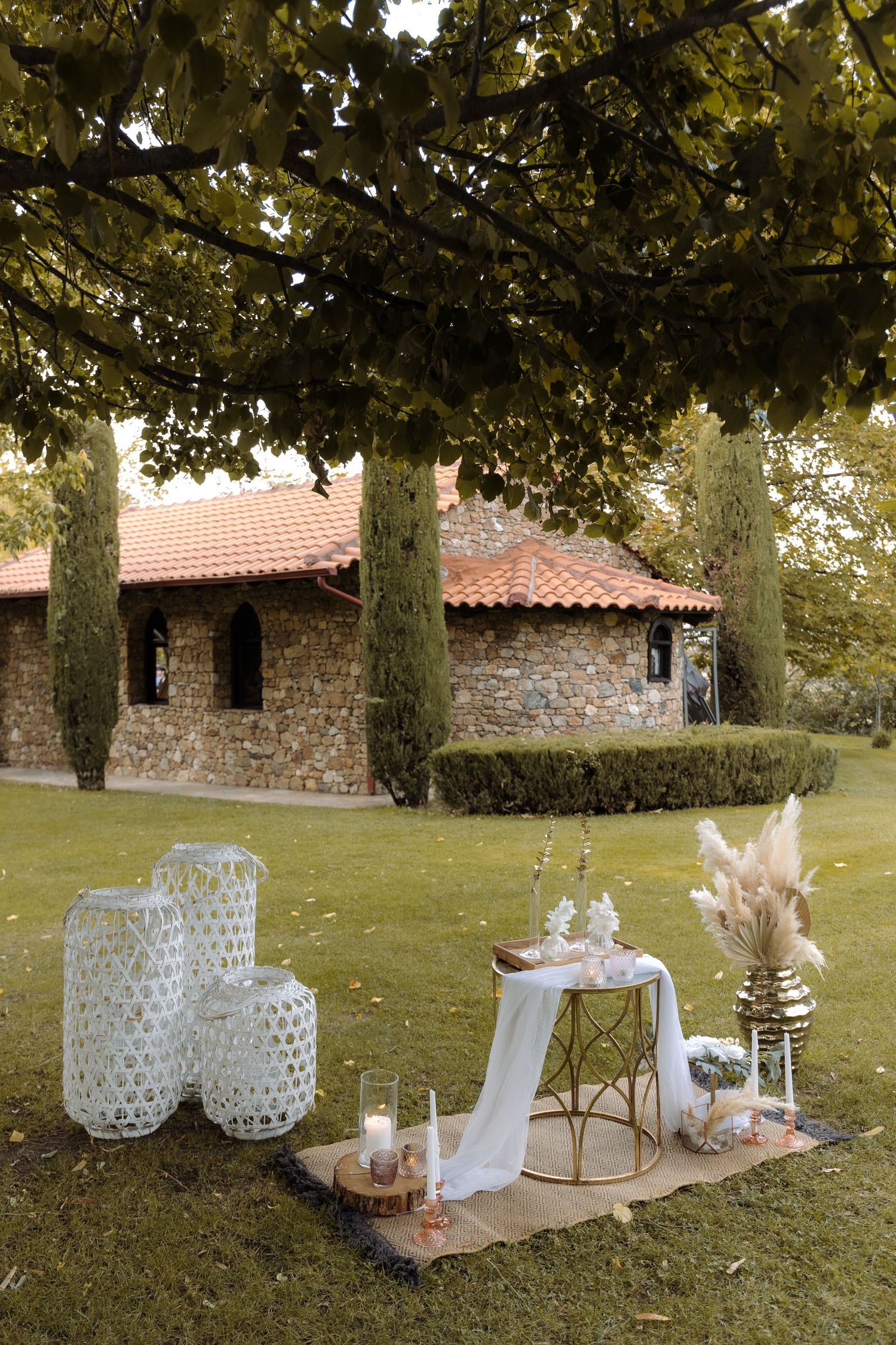 Romantic outdoor wedding setup with white lanterns and decor beneath cypress trees near a rustic stone building.