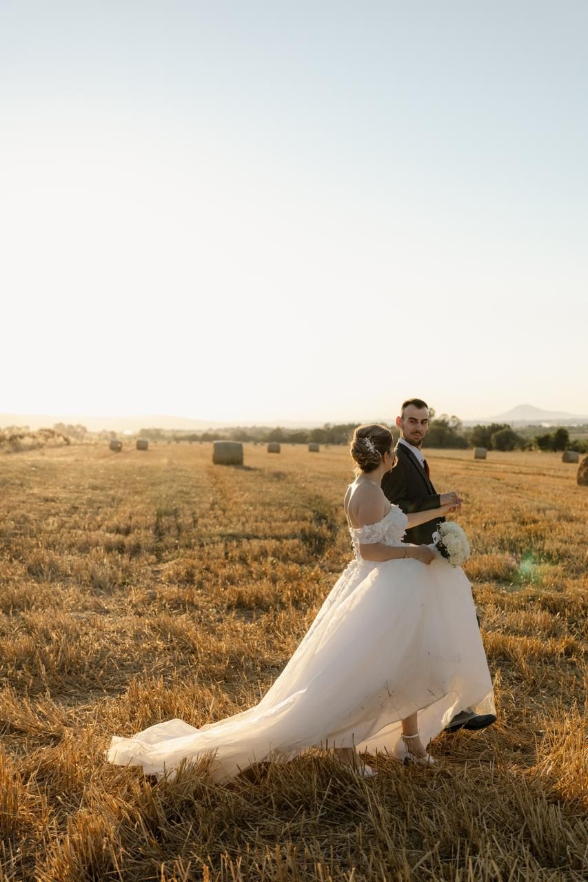Bride and groom dancing in golden wheat field at sunset, with hay bales and rolling hills in the background.