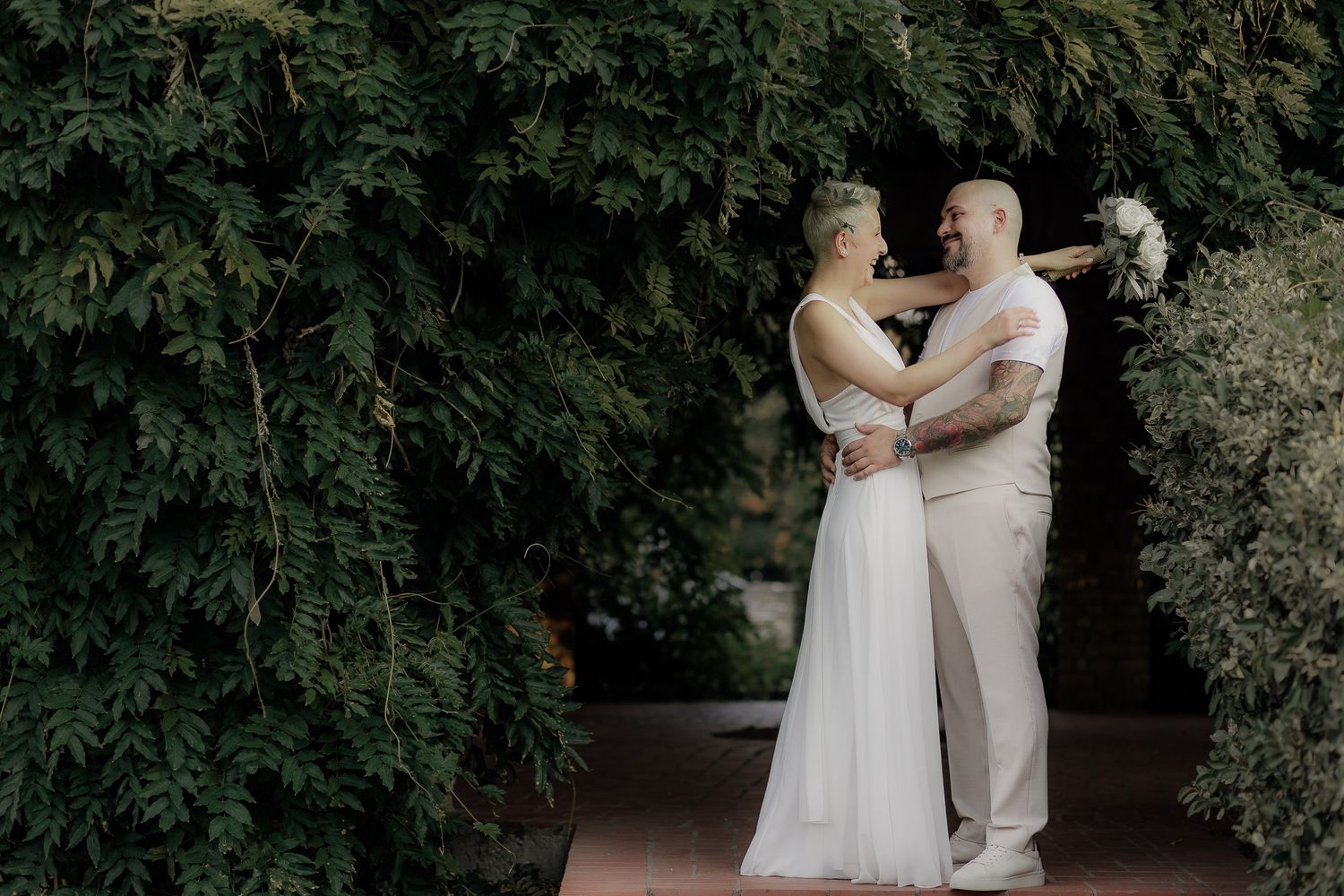 A romantic wedding couple embraces in a lush garden setting with green foliage backdrop.