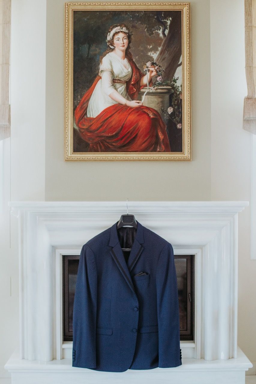 Navy blue groom's suit jacket displayed beneath vintage portrait painting on white fireplace mantel.