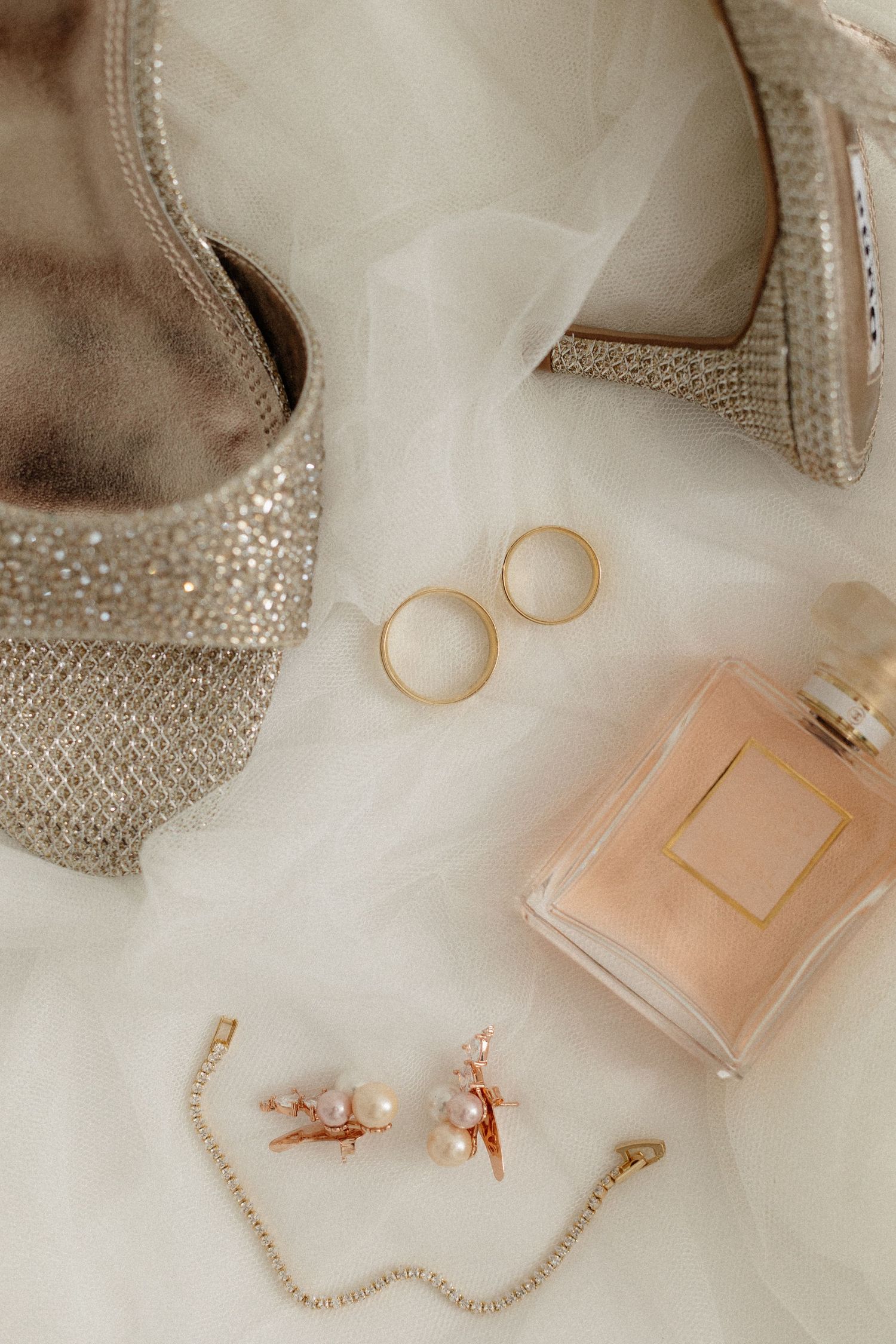 Wedding accessories including sparkly shoes, gold rings, perfume and jewelry on white fabric.