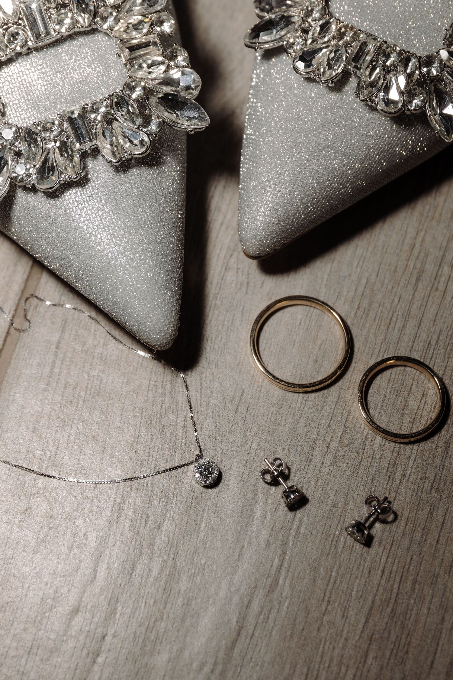 Wedding accessories including silver shoes, wedding rings and delicate jewelry on wooden surface.