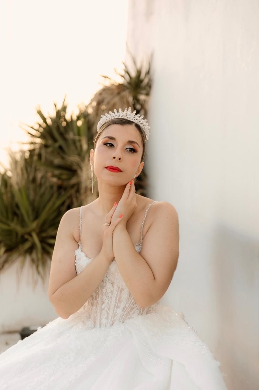 Elegant bride in white lace gown and crystal tiara poses gracefully with hands near face against white wall.