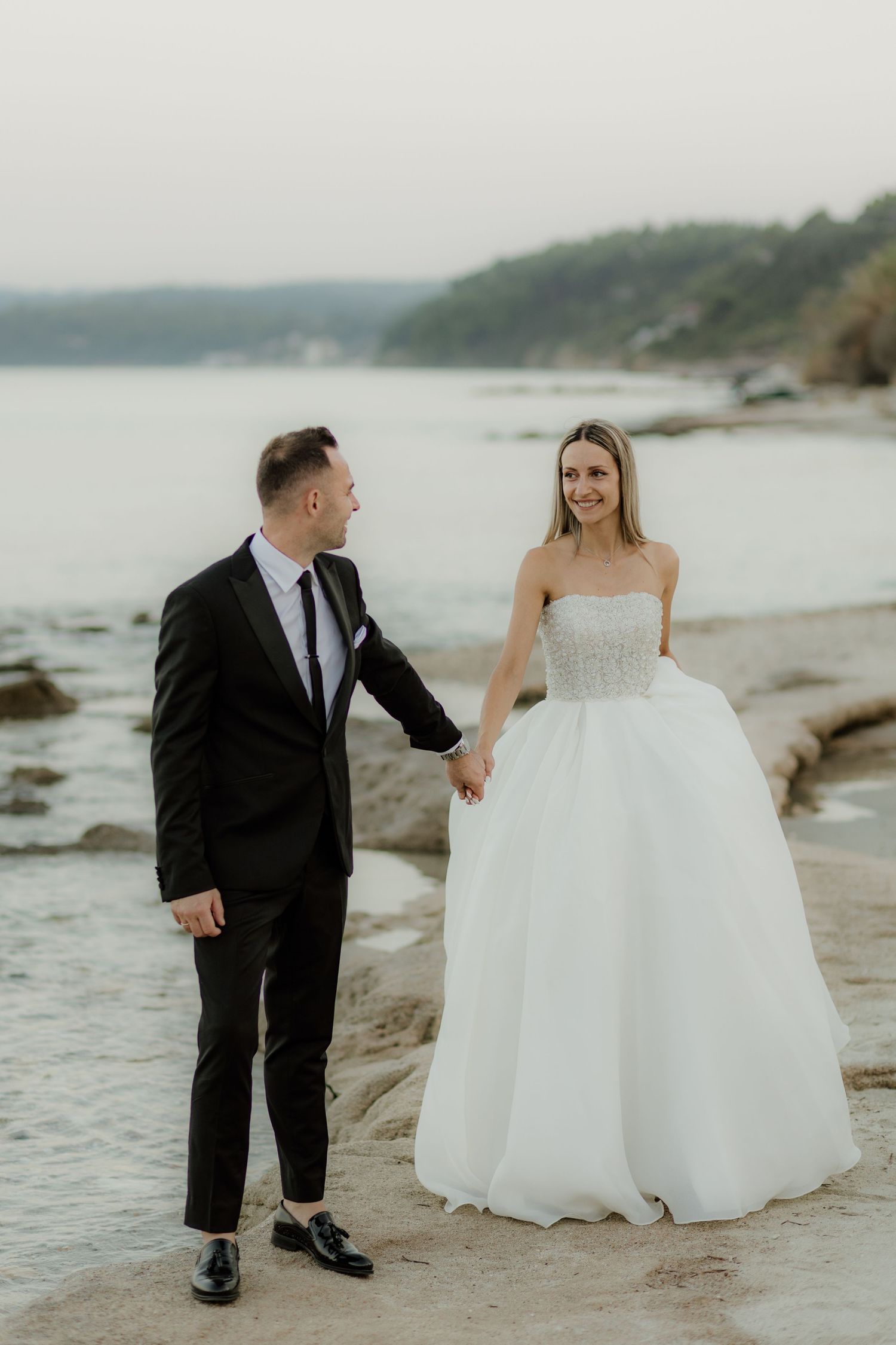 A couple walking along a rocky beach shoreline with the bride wearing a flowing white gown.