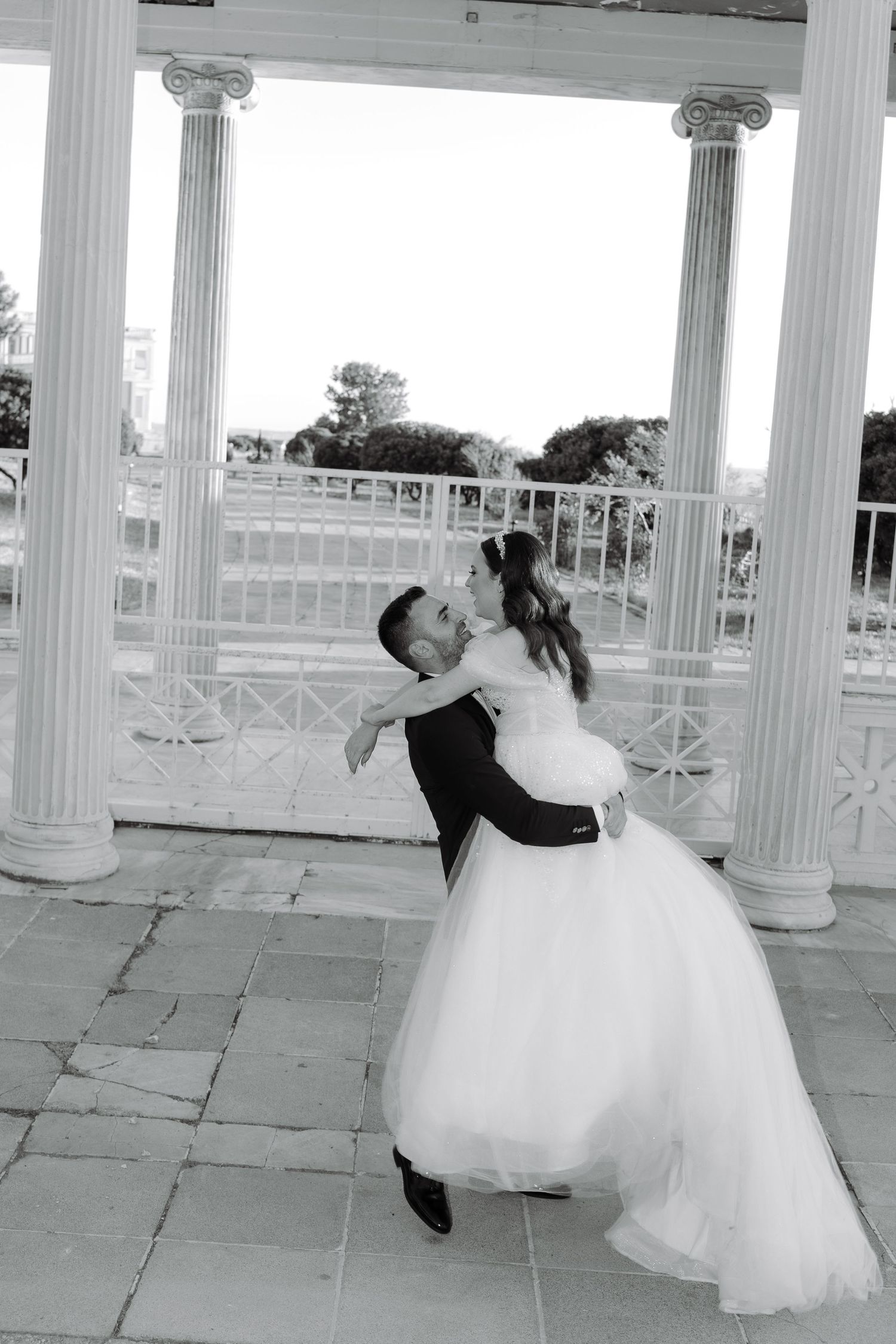 Romantic black and white photo of newlyweds dancing under a classical columned pavilion.