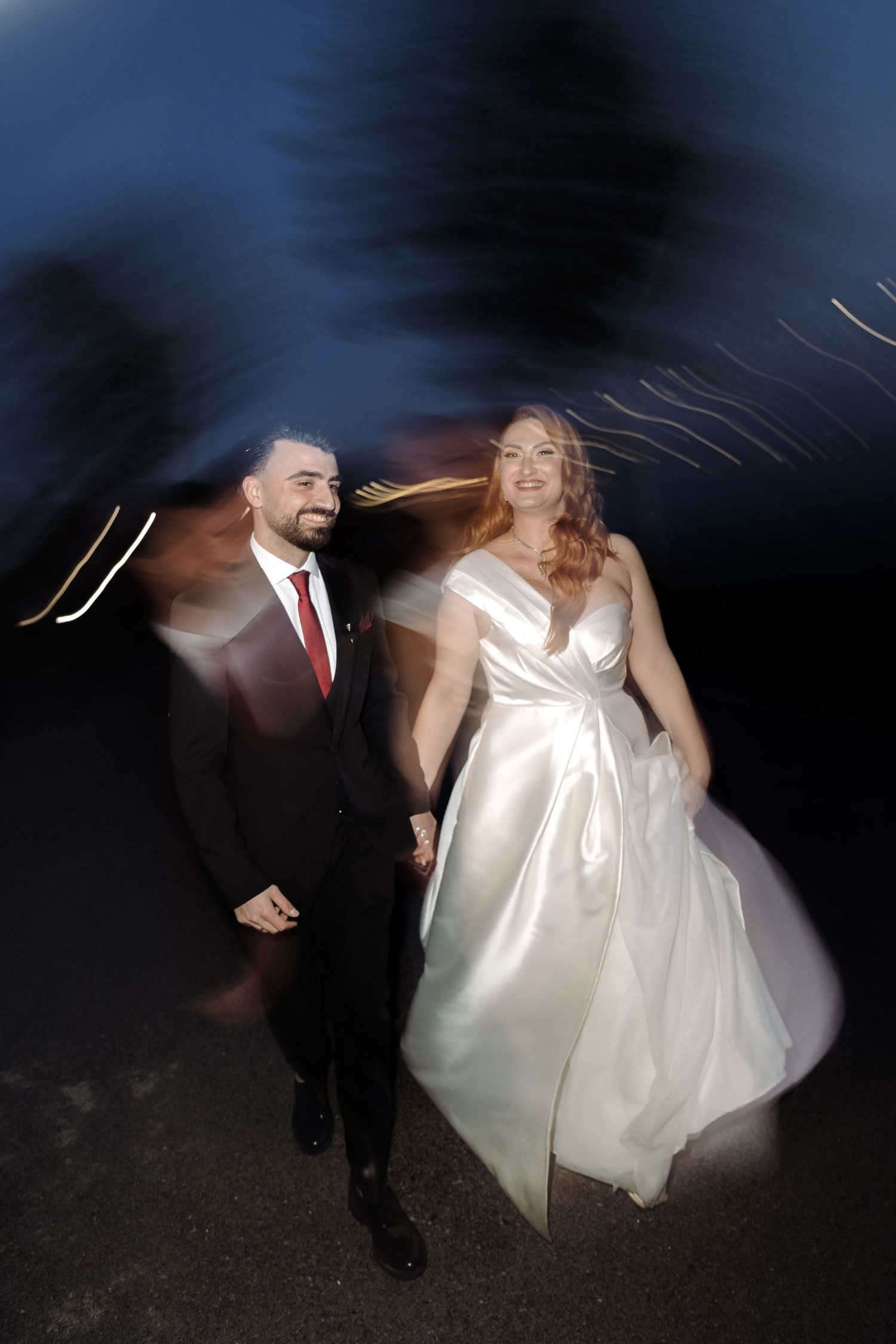 Bride in white gown and groom in black suit hold hands walking at night with light streaks in background.