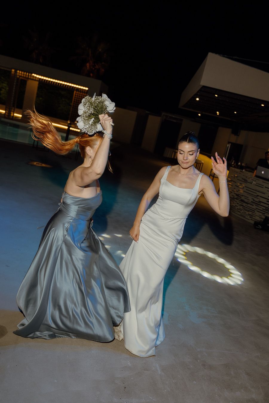 Bride and bridesmaid dancing outdoors at night, wearing elegant white and silver gowns, with bouquet raised.