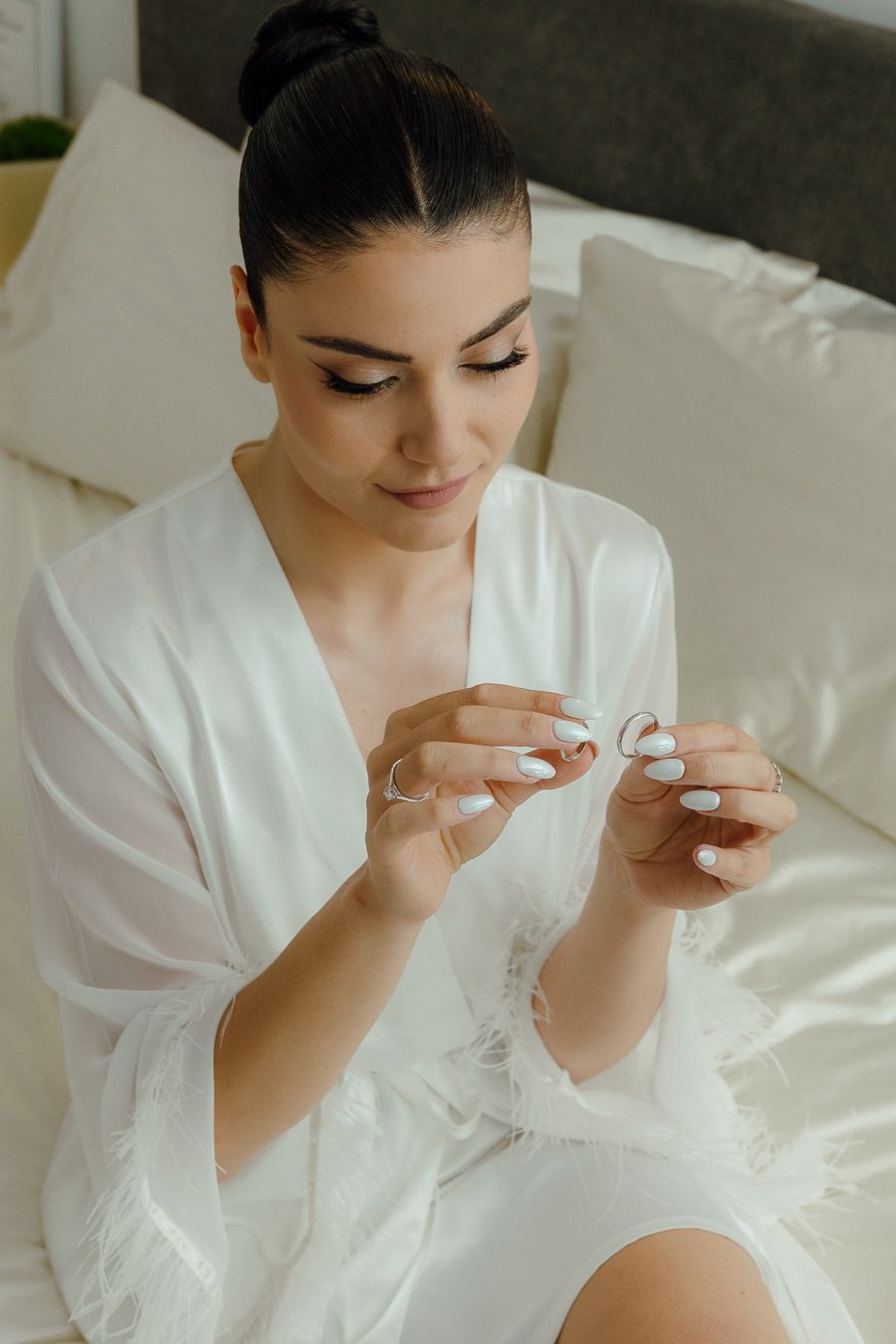 Bride in white satin robe sitting on bed examining wedding rings with elegant updo and manicured nails.