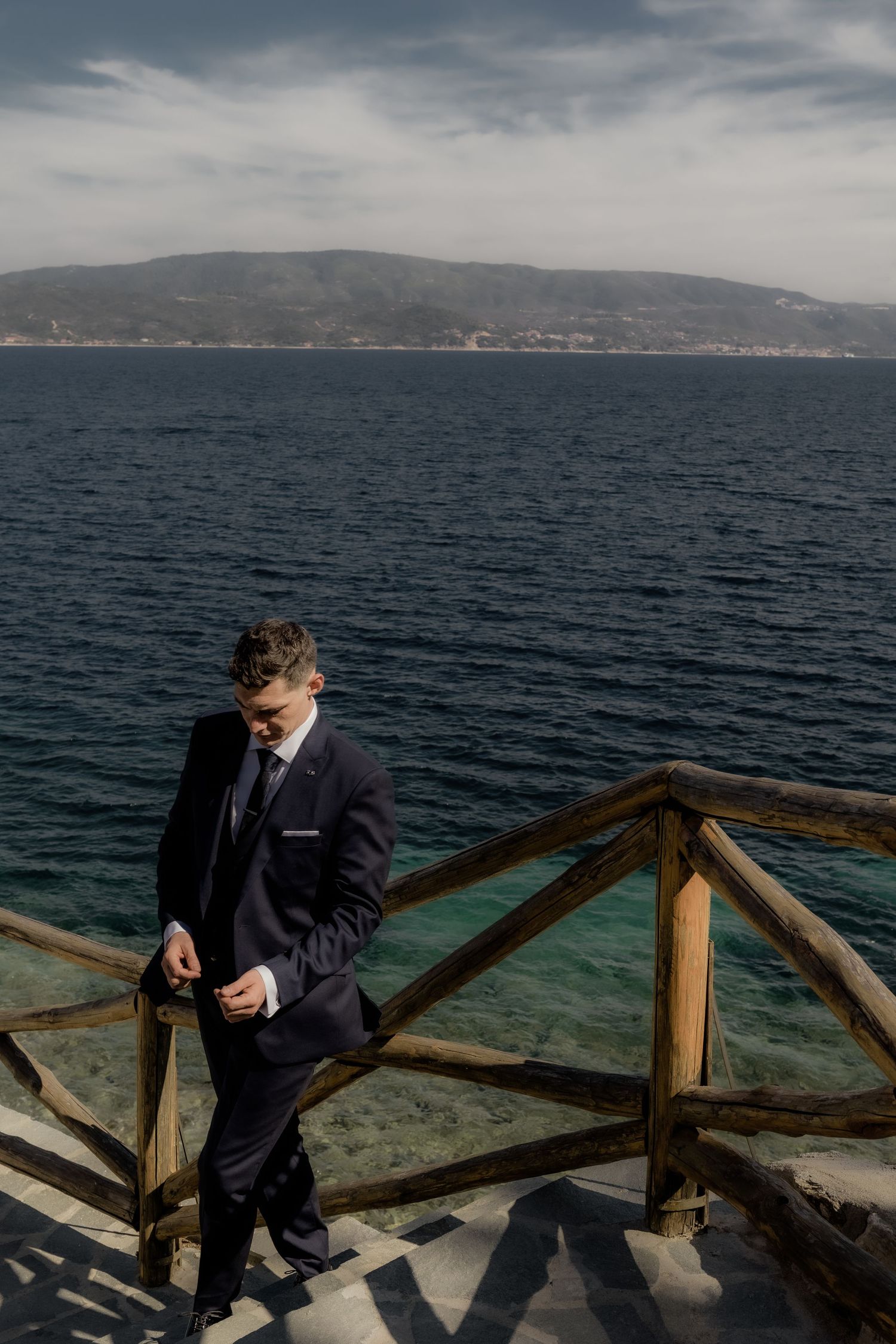 Person in dark suit standing on wooden deck overlooking dramatic ocean view.