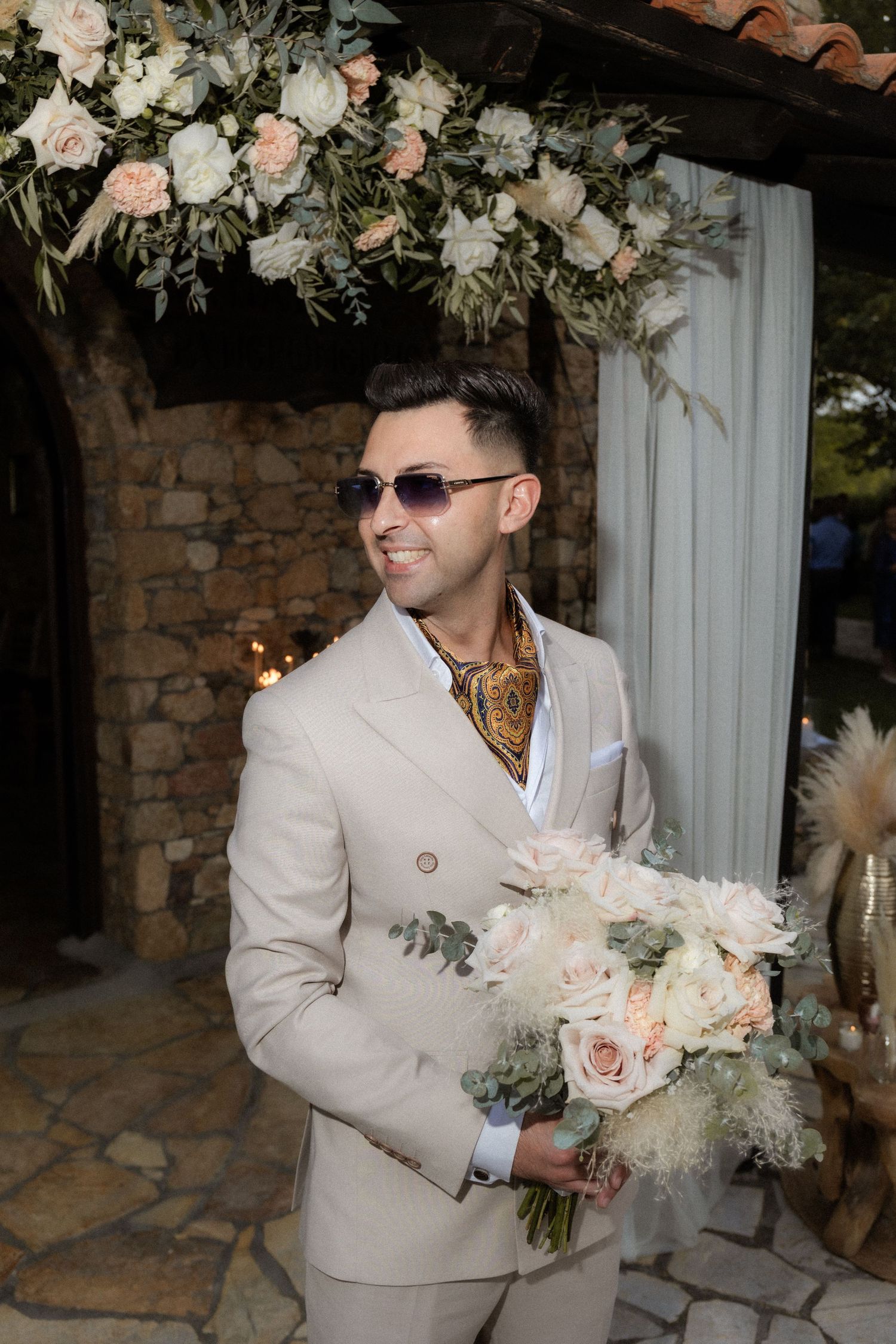 Wedding attendant in beige suit holding delicate pink and white floral bouquet against stone wall backdrop.